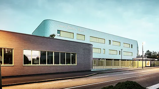 Abendansicht des Collège Léontine Dolivet in Cesson-Sévigné. Im Vordergrund verläuft eine Straße mit klarer Markierung, daneben ein flacher Gebäudeteil mit großen Glasfenstern. Dahinter erhebt sich der Hauptbau mit geschwungener Aluminiumfassade aus PREFA Wandrauten in Silbermetallic, die im sanften Licht changiert. Mehrere rechteckige Fenster und ein heller Zaun strukturieren die Ansicht. Die Szene ist von Straßenlaternen beleuchtet, der Himmel zeigt ein kühles Blau – ein moderner, eleganter Blickfang.