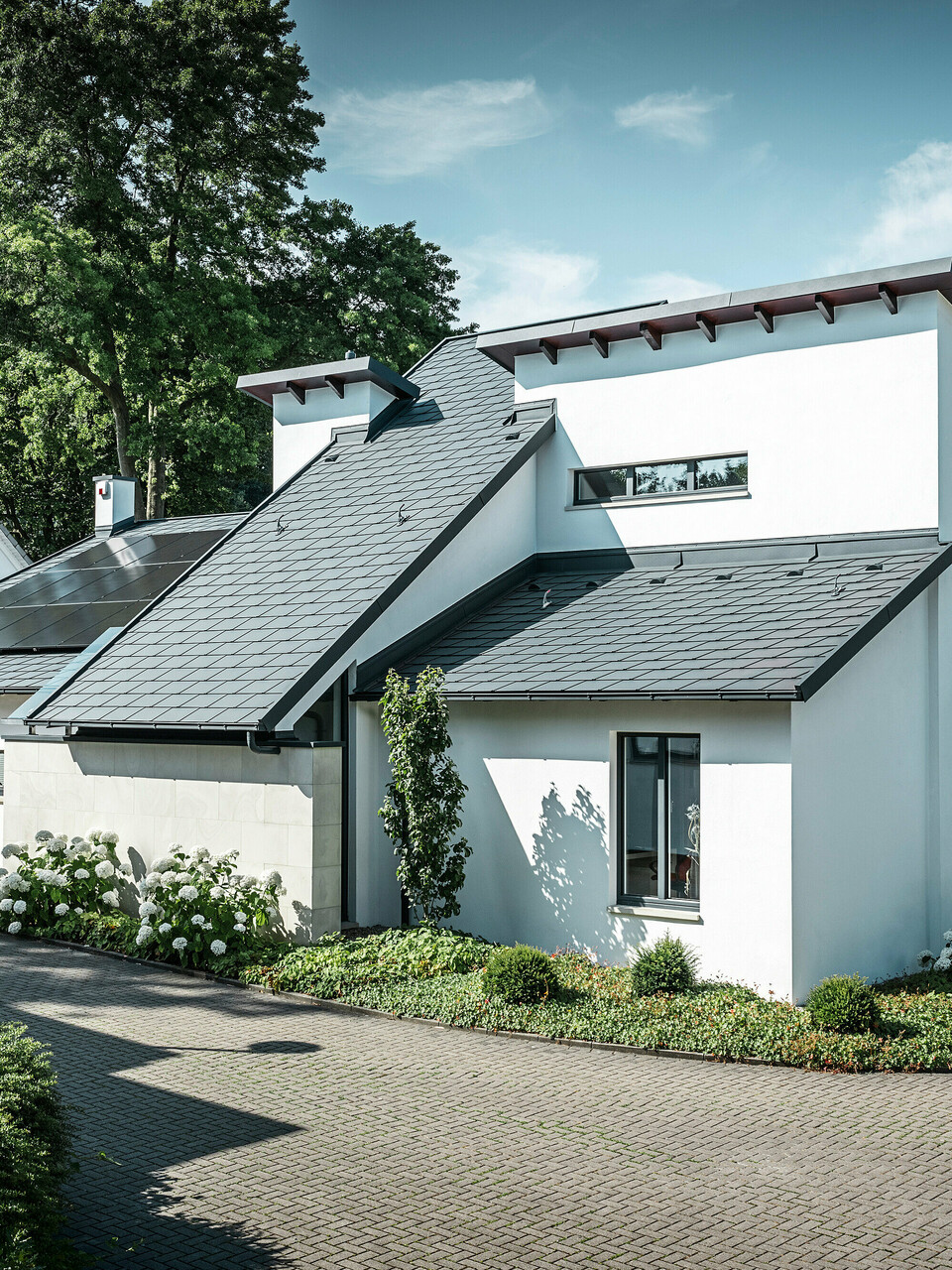 Modernes Einfamilienhaus mit weißer Putzfassade und dunklem PREFA Schrägdach aus Aluminium. Auf dem Dach sind integrierte Photovoltaikmodule und eine Aluminium-Dachentwässerung in Anthrazit zu sehen. Das Gebäude liegt in einer gepflegten Wohnsiedlung mit bepflanzten Vorgärten, gepflasterter Zufahrt und umliegenden Einfamilienhäusern.