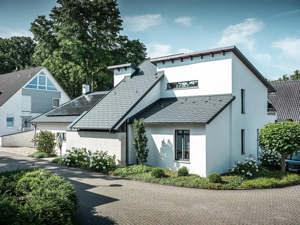 Modern detached house with white plaster façade and dark PREFA aluminium pitched roof. Integrated photovoltaic modules and anthracite aluminium roof drainage can be seen on the roof. The building is located in a well-maintained residential area with planted front gardens, paved driveways and surrounding detached houses.