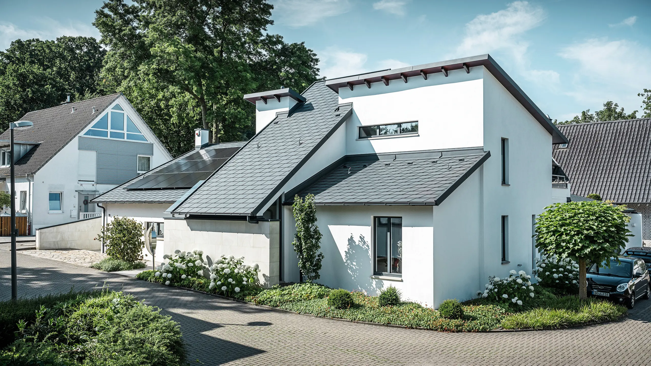 Modern detached house with white plaster façade and dark PREFA aluminium pitched roof. Integrated photovoltaic modules and anthracite aluminium roof drainage can be seen on the roof. The building is located in a well-maintained residential area with planted front gardens, paved driveways and surrounding detached houses.