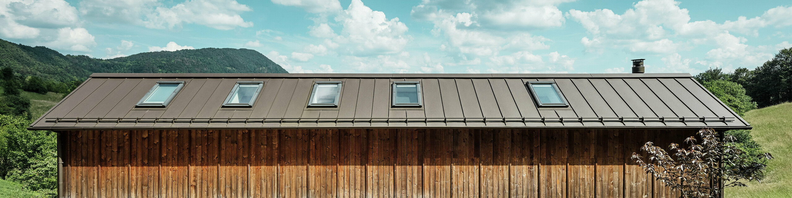Dieses moderne Wohnhaus in Hanglage besticht durch seine minimalistische Holzfassade und das langlebige PREFALZ-Aluminiumdach in der Farbe P.10 Braun. Die Stehfalz-Dachdeckung überzeugt durch ihre hohe Wetterbeständigkeit und Langlebigkeit sowie durch das harmonische Zusammenspiel mit der natürlichen Umgebung. Integrierte Dachfenster und eine klare Linienführung betonen die zeitlose Architektur. Auf dem Bild ist der Eingangsbereich des kleinen Einfamilienhauses zu erkennen.