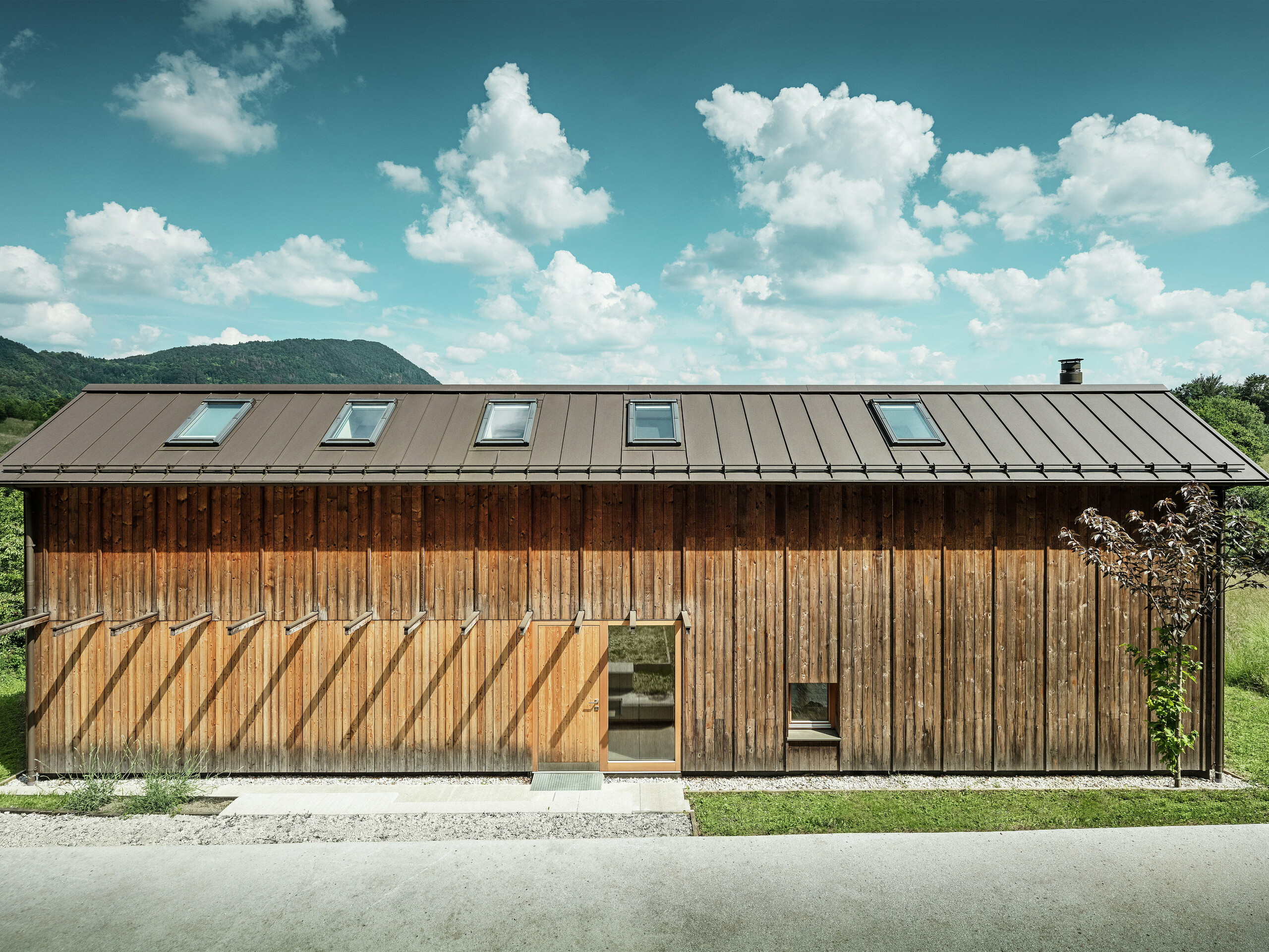 Dieses moderne Wohnhaus in Hanglage besticht durch seine minimalistische Holzfassade und das langlebige PREFALZ-Aluminiumdach in der Farbe P.10 Braun. Die Stehfalz-Dachdeckung überzeugt durch ihre hohe Wetterbeständigkeit und Langlebigkeit sowie durch das harmonische Zusammenspiel mit der natürlichen Umgebung. Integrierte Dachfenster und eine klare Linienführung betonen die zeitlose Architektur. Auf dem Bild ist der Eingangsbereich des kleinen Einfamilienhauses zu erkennen.