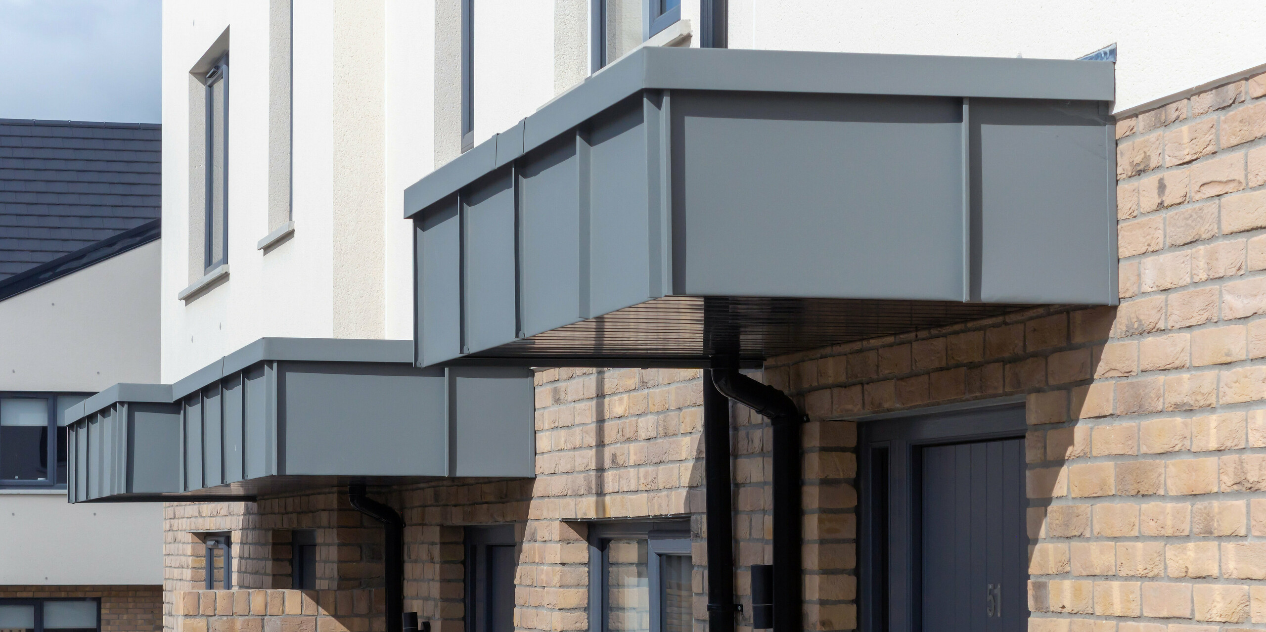 Das Bild zeigt die markanten Eingangsvordächer der Reihenhäuser in Churchfields, die vollständig mit PREFALZ in P.10 Hellgrau verkleidet sind. Die klaren, geradlinigen Aluminiumflächen setzen einen modernen Akzent über dem warmen Backsteinmauerwerk und verleihen der Anlage ein einheitliches, hochwertiges Erscheinungsbild. Die robuste PREFA Oberfläche ist witterungsbeständig, farbstabil und nahezu wartungsfrei – ideal für langlebige Wohnbauten. Die Detailaufnahme betont die präzise Verarbeitung und die harmonische Integration der Aluminiumverkleidung in die Architektur.