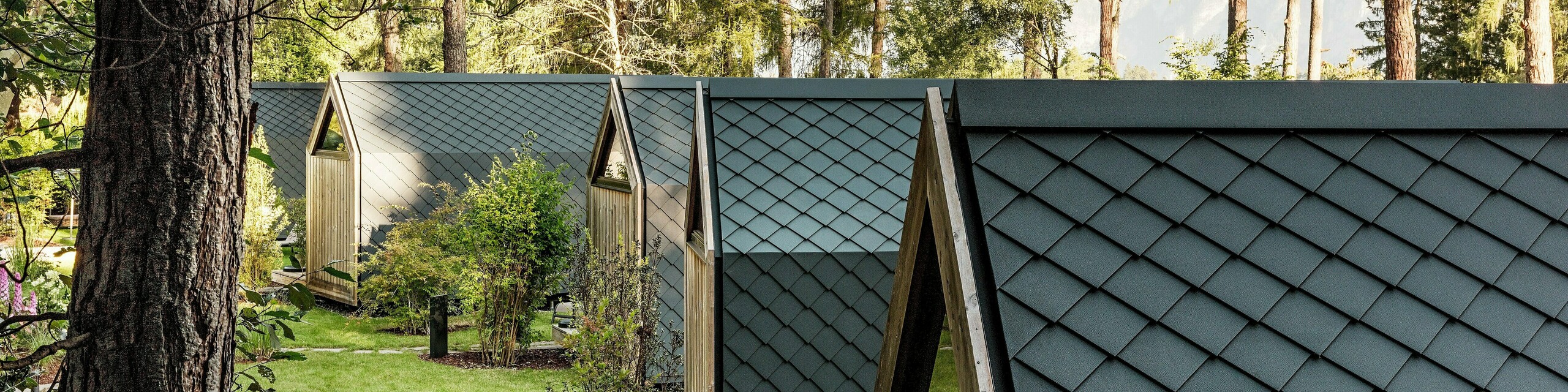 Mehrere minimalistische Tiny Houses des Secret Koi Garden reihen sich harmonisch entlang einer grünen Gartenachse am Natterer See. Die Dächer und Fassaden sind vollständig mit kleinteiligen PREFA Dach‑ und Wandrauten aus Aluminium in P.10 Anthrazit verkleidet und bilden eine gleichmäßige, fischhautartige Oberfläche. Umgeben von Bäumen und Sträuchern verbinden die langlebigen, witterungsbeständigen PREFA Aluminiumprodukte präzise Architektur mit Ruhe, Natur und hochwertigem Glamping‑Design.