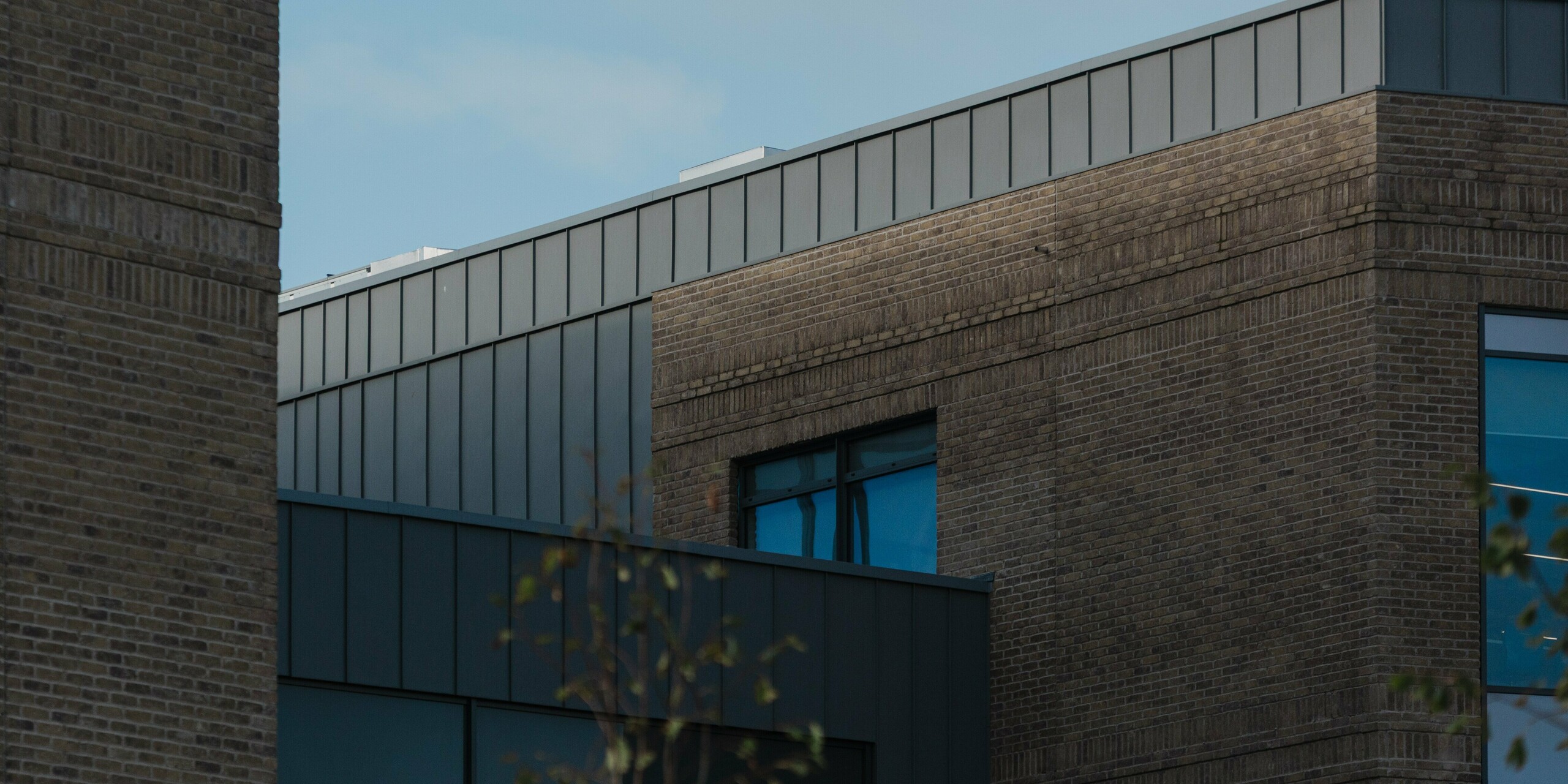 A close-up architectural view of the Northern Regional College in Ballymena highlights the upper façade edge finished with vertical PREFALZ standing seam aluminium panels in patina grey. The crisp aluminium band creates a strong contrast to the textured brickwork and recessed windows beneath. Set against a blue sky, the smooth metal surface subtly reflects daylight and reinforces the building’s clean geometry. PREFA aluminium delivers durability, precise detailing and a timeless, low-maintenance façade solution for contemporary educational architecture.