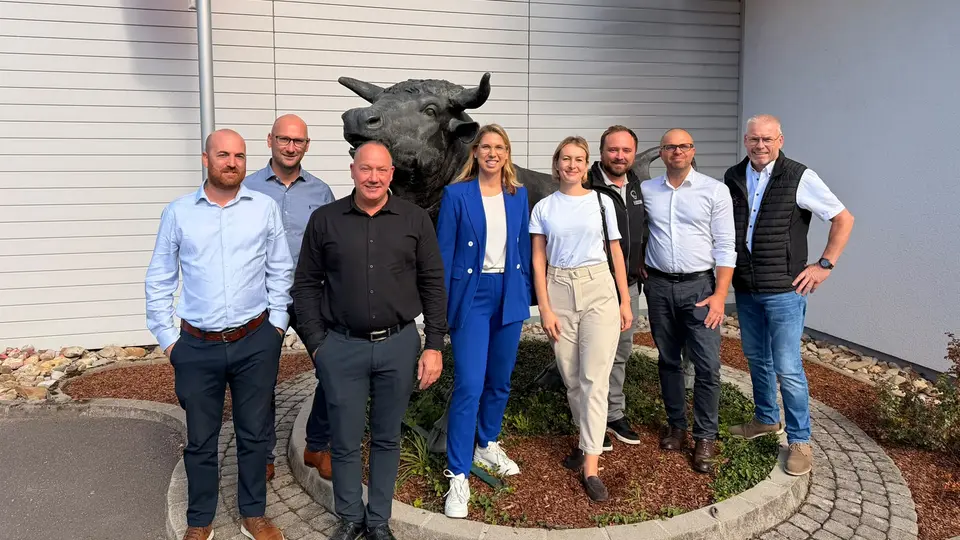A group photo of eight people outdoors in front of a company building, with a large sculpture of a bull in the background.