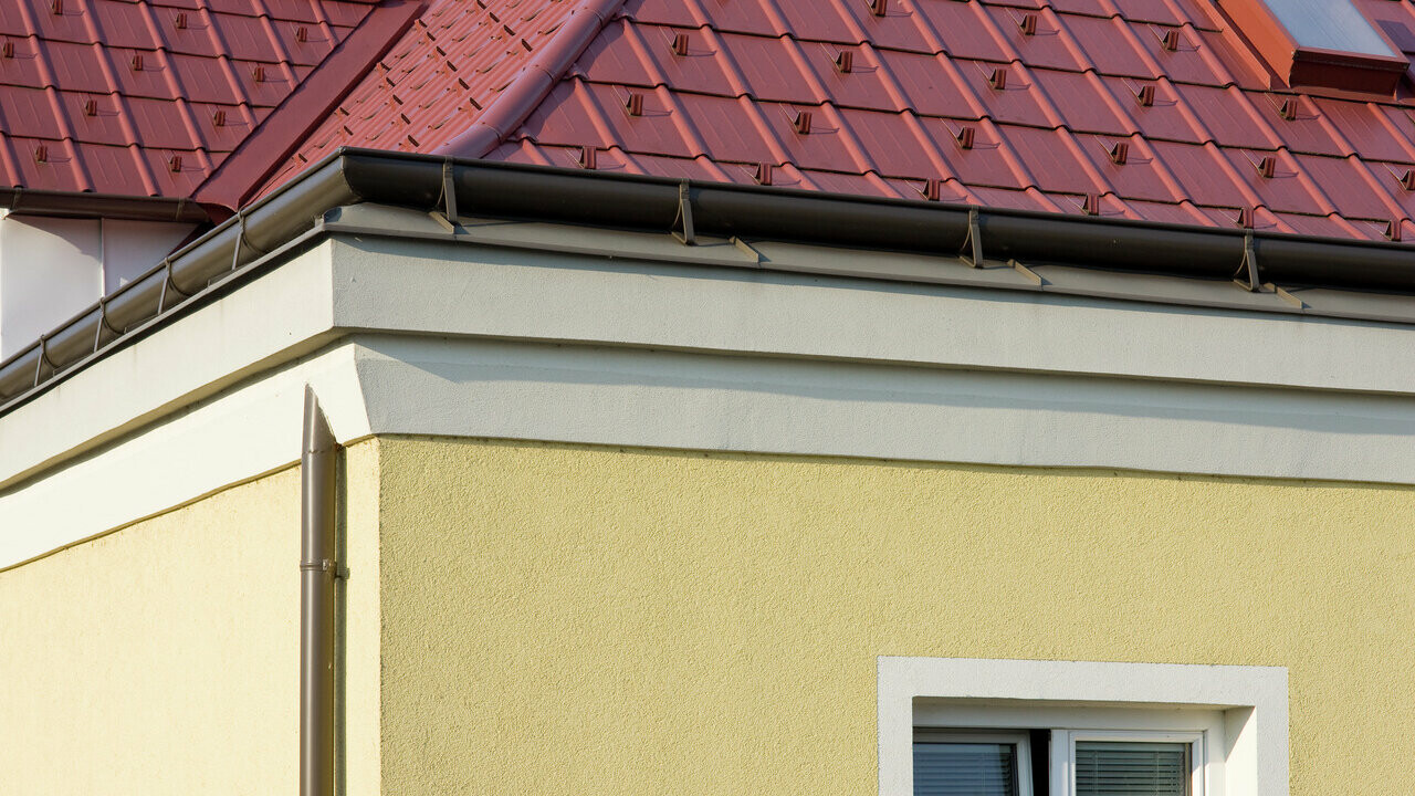 Corner of house with yellow plaster façade and brown PREFA on-roof gutter with red PREFA roof tile roof