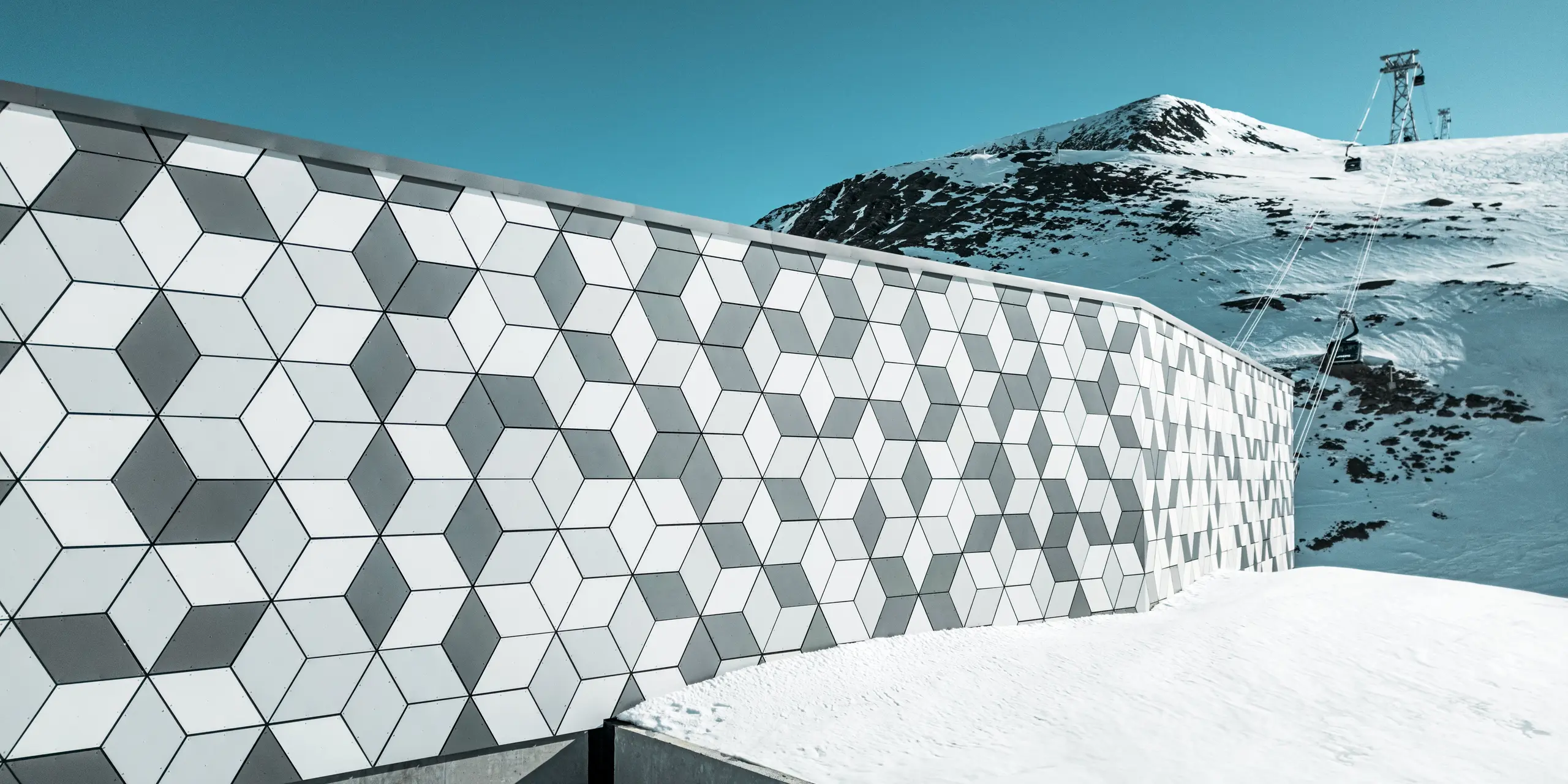 Die Fassade der Seilbahnstation in Les Deux Alpes ist ein Meisterwerk moderner Architektur. Sie besteht aus PREFABOND Aluminium Verbundplatten, die in einem auffälligen Rautenmuster aus Weiß und Grau angeordnet sind. Die geometrische Gestaltung fügt sich harmonisch in die schneebedeckte Berglandschaft ein und bietet dank der robusten und wetterbeständigen Materialien lang anhaltenden Schutz vor extremen Witterungsbedingungen. Der Blick vorbei an der Fassade zeigt den Weg der Gondel hinauf zum Berg.