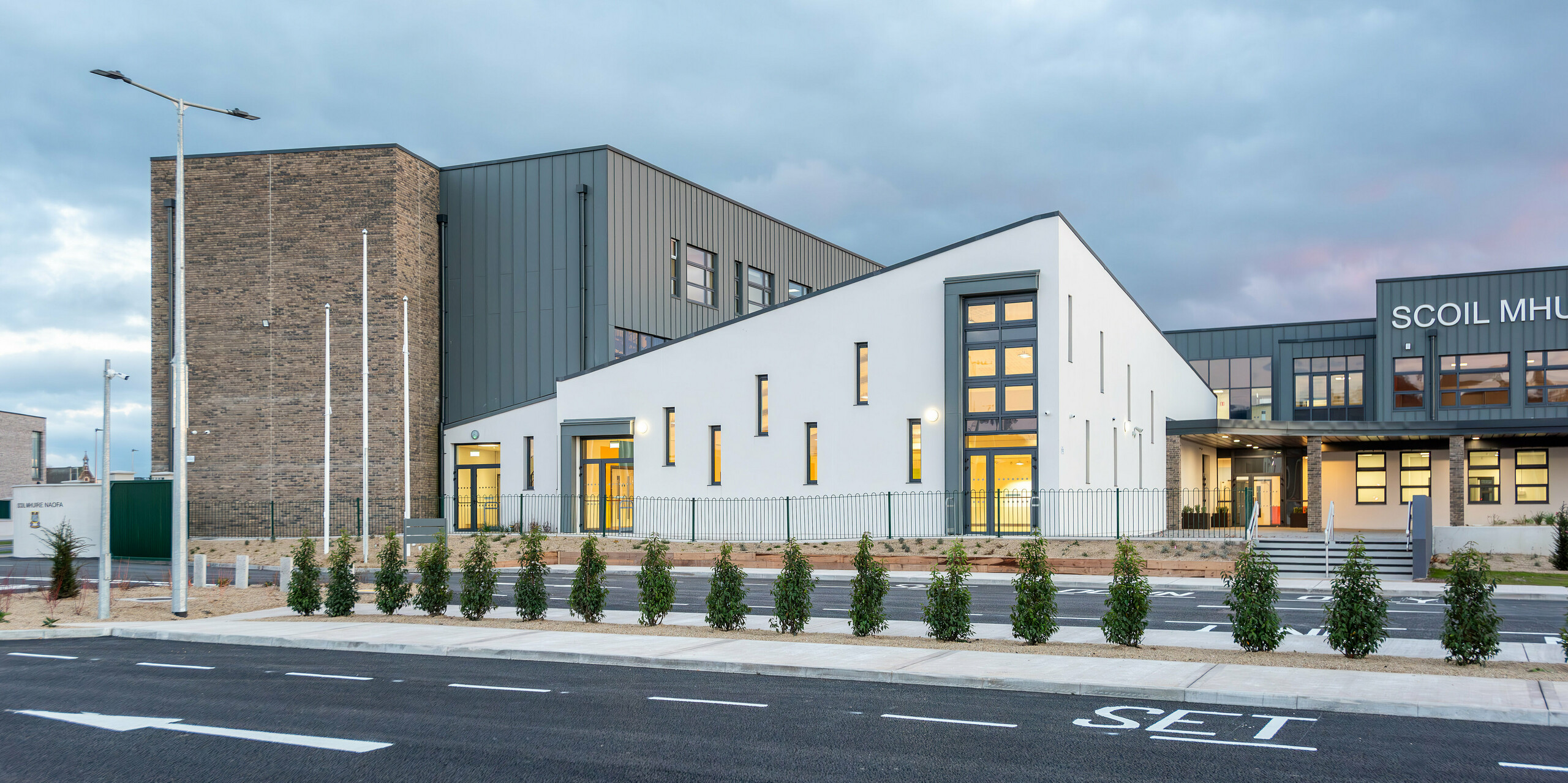 The modern Scoil Mhuire Naofa school in Carrigtwohill, County Cork, Republic of Ireland, with its elegant PREFALZ aluminium facade in P.10 light grey. The bright, minimalist design combines clean lines with high-quality standing seam cladding, while the surrounding landscape and the new driveway emphasise the modern character of the educational institution. The illuminated interiors draw attention to the generous windows.