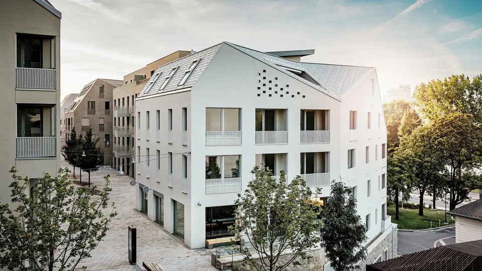Modernes Wohnquartier Vydrica in Bratislava mit klarer Architektursprache und ruhiger Gassenatmosphäre: Die steil geneigten Satteldächer mit markanten Dachfenstern sind mit PREFA Aluminium Dachschindeln in P.10 Prefaweiß, Bronze und P.10 Sandbraun gedeckt und bilden einen eleganten Eyecatcher im Sonnenlicht. Die präzise ausgeführten Linien und homogenen Flächen unterstreichen die hochwertige Gestaltung. PREFA Aluminium überzeugt durch Langlebigkeit, geringes Gewicht und hohe Witterungsbeständigkeit – ideal für komplexe Dachformen und eine nachhaltige, wartungsarme Gebäudehülle.