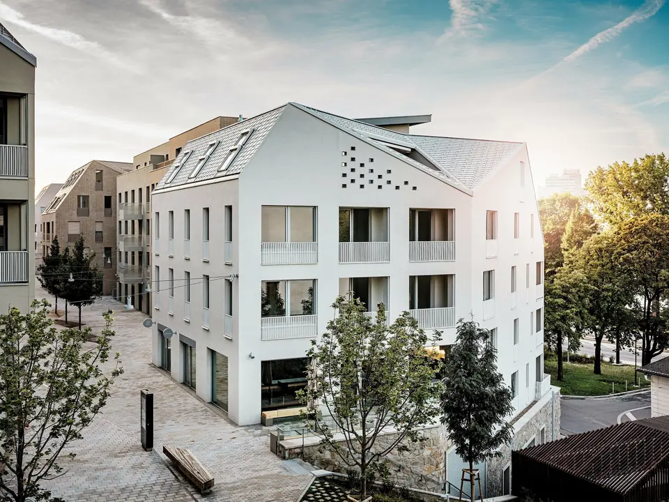 Modernes Wohnquartier Vydrica in Bratislava mit klarer Architektursprache und ruhiger Gassenatmosphäre: Die steil geneigten Satteldächer mit markanten Dachfenstern sind mit PREFA Aluminium Dachschindeln in P.10 Prefaweiß, Bronze und P.10 Sandbraun gedeckt und bilden einen eleganten Eyecatcher im Sonnenlicht. Die präzise ausgeführten Linien und homogenen Flächen unterstreichen die hochwertige Gestaltung. PREFA Aluminium überzeugt durch Langlebigkeit, geringes Gewicht und hohe Witterungsbeständigkeit – ideal für komplexe Dachformen und eine nachhaltige, wartungsarme Gebäudehülle.