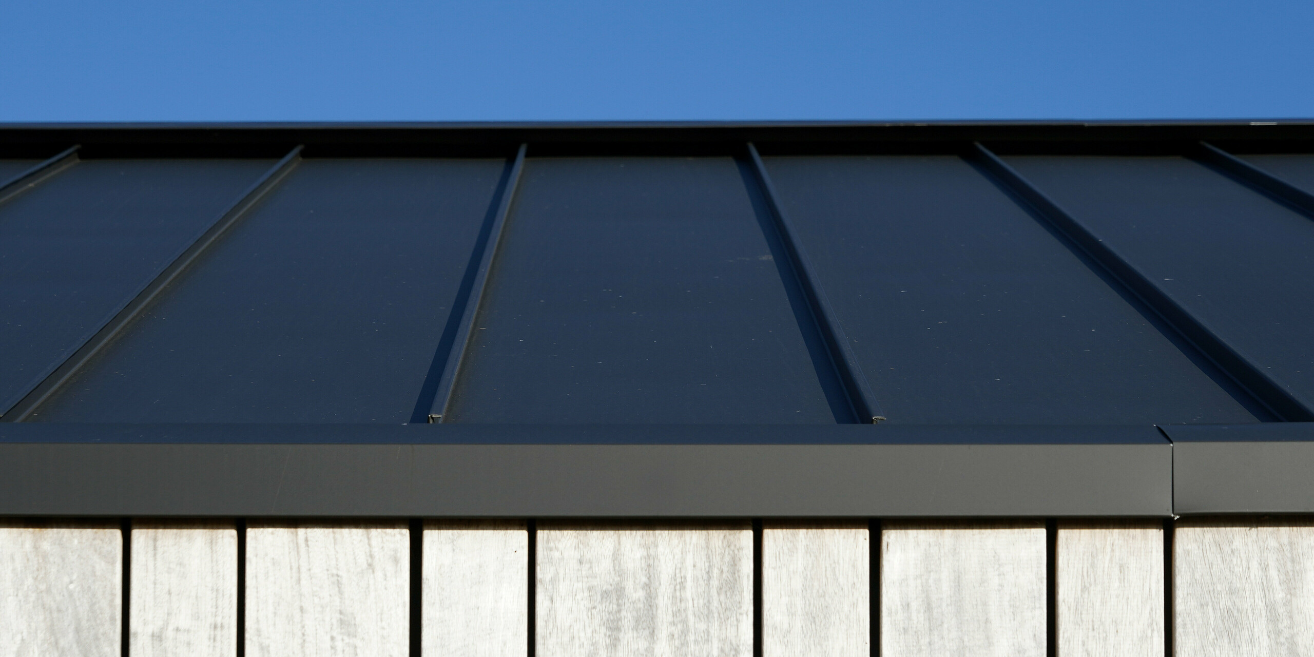 Detailaufnahme eines modernen Einfamilienhauses in Uithoorn, Niederlande, mit PREFALZ Winkelstehfalz-Eindeckung in Schwarzgrau. Das Bild zeigt die präzise Verarbeitung des hochwertigen Blechs von PREFA, kombiniert mit natürlichen Holzelementen an der Fassade. Die klare Linienführung des robusten Aluminiumdachs unterstreicht die langlebige und wetterbeständige Bauweise.