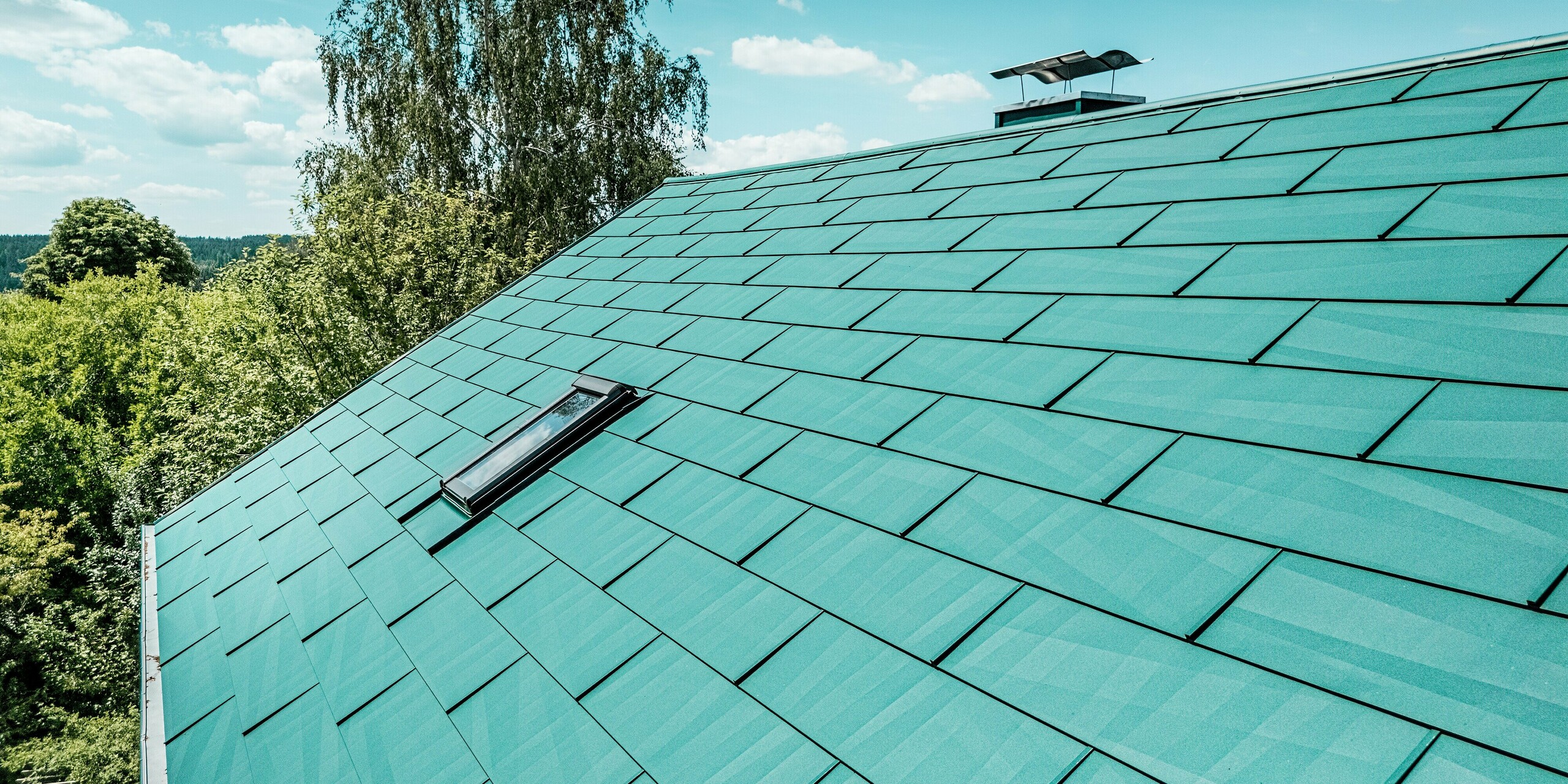 Detailaufnahme des sanierten Dachs eines Einfamilienhauses in Schwarzenbach-Martinlamitz mit PREFA Dachpaneel FX.12 in P.10 Moosgrün. Das Bild zeigt die strukturierte Aluminiumoberfläche in moderner, unregelmäßiger Kantung, die das Sonnenlicht stilvoll reflektiert. Das markante Dachfenster und der elegant verkleidete Kamin setzen Akzente. Die langlebigen PREFA Paneele bieten optimalen Schutz vor Witterungseinflüssen, sind wartungsfrei, sturmsicher und besonders leicht – ideal für hochwertige Dachsanierungen mit architektonischem Anspruch.