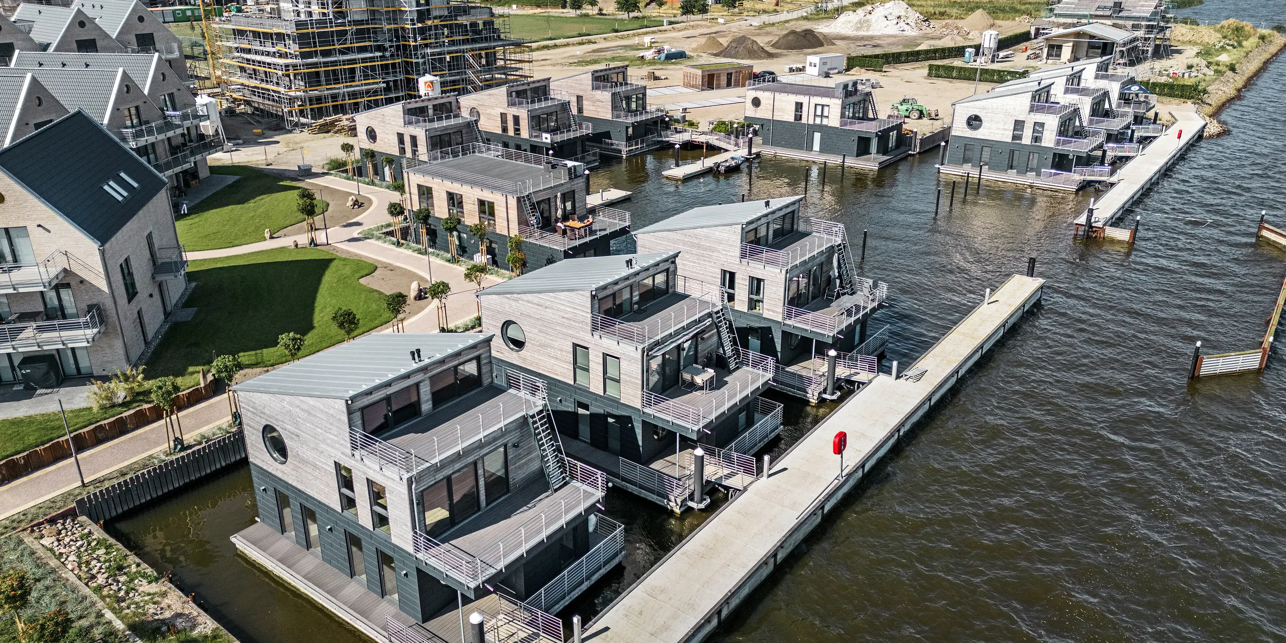 Luftaufnahme der innovativen Wasserhaus-Siedlung im Pionierhafen Schleswig, eingebettet in einer maritimen Umgebung und angrenzender Bebauung im Aufbau. Die elf schwimmenden Domizile, charakteristisch verkleidet mit PREFA Siding.X in P.10 Anthrazit, harmonieren mit den Holzelementen an der Fassade. Die PREFA Aluminiumprodukte stehen für ihre Beständigkeit gegenüber der direkten Wassernähe. Die architektonische Symbiose von funktionaler Eleganz und natürlicher Ästhetik entlang des Ufers unterstreicht die Exklusivität dieser Wohnanlage, die primär als Feriendestination dient.