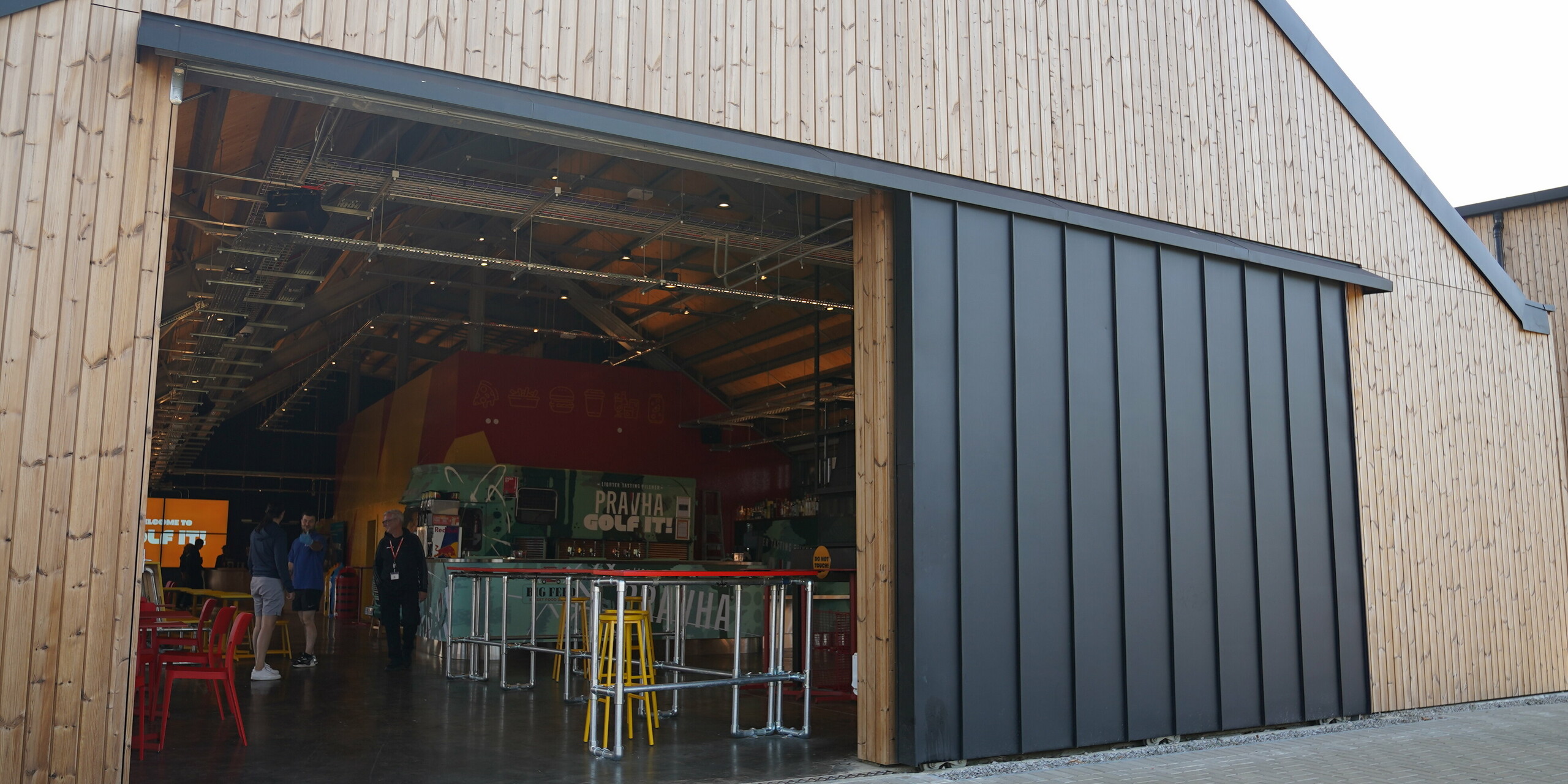 A view of the open entrance area of the "Golf it!" facility in Glasgow, Scotland. The construction demonstrates the versatile use of PREFALZ in P.10 black. The large sliding door is framed by a wooden façade on the building. Inside, a lively atmosphere with colourful seating and bar tables invites people to linger. The flexible aluminium was used in this project on around 2,000 square metres of roof area, on the large sliding doors and parts of the façade, creating a harmonious appearance.