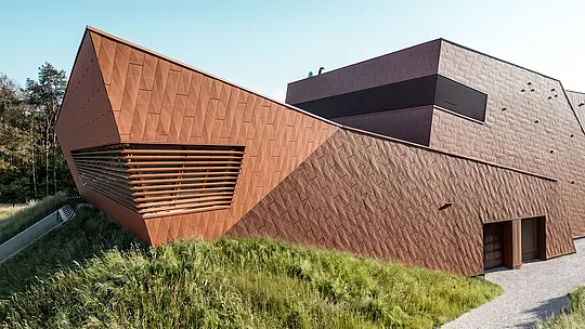 Ausdrucksstarkes Trinkwasserpumpwerk WML in Lottum mit dynamisch gefaltetem Baukörper und markanter Gebäudespitze als visueller Eyecatcher. Die PREFA Siding.X Aluminiumfassade in warmer Rostoptik beeindruckt durch ihre facettierte, lebendige Struktur und das präzise Spiel von Licht und Schatten. Horizontale Lamellen betonen die Tiefe der Architektur, während sich das Gebäude harmonisch in die natürliche Umgebung einfügt. Die leichten, langlebigen und recycelbaren PREFA Aluminium-Paneele stehen für höchste Witterungsbeständigkeit sowie eine nachhaltige, wartungsarme Gebäudehülle.