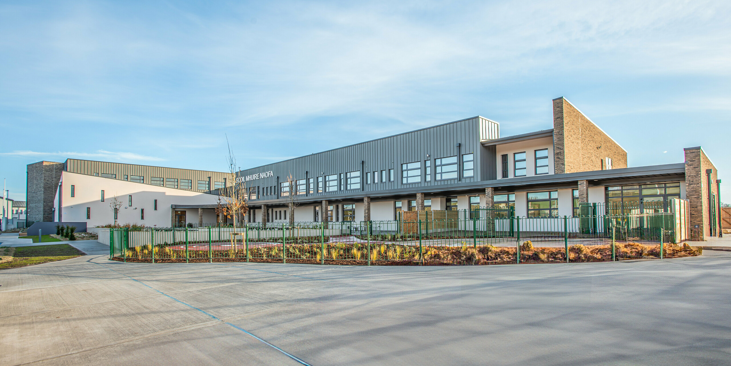 A wide-angle view of Scoil Mhuire Naofa in Carrigtwohill, Republic of Ireland. The school building combines modern architecture with a PREFALZ aluminium façade in P.10 light grey. The durable and weather-resistant standing seam roof emphasises the functional elegance, while the well-kept grounds underscore the modern character of the school.