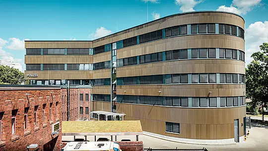 Dynamische Außenansicht des erweiterten Polizeikommissariats 43 in Hamburg: Der geschwungene Neubau mit rund 1.100 m² PREFA Aluminiumfassade in mittelbronze eloxierten Strangpressprofilen „Zacke“ setzt als Eyecatcher einen markanten Kontrast zum Backsteinbestand. Die fein strukturierte Oberfläche spielt lebendig mit Licht und Schatten und verleiht der Fassade Tiefe und Präzision. Dank hoher Korrosionsbeständigkeit, Langlebigkeit und wartungsarmer Eigenschaften stehen PREFA Aluminiumlösungen für nachhaltige, funktionale und architektonisch anspruchsvolle Bauqualität.