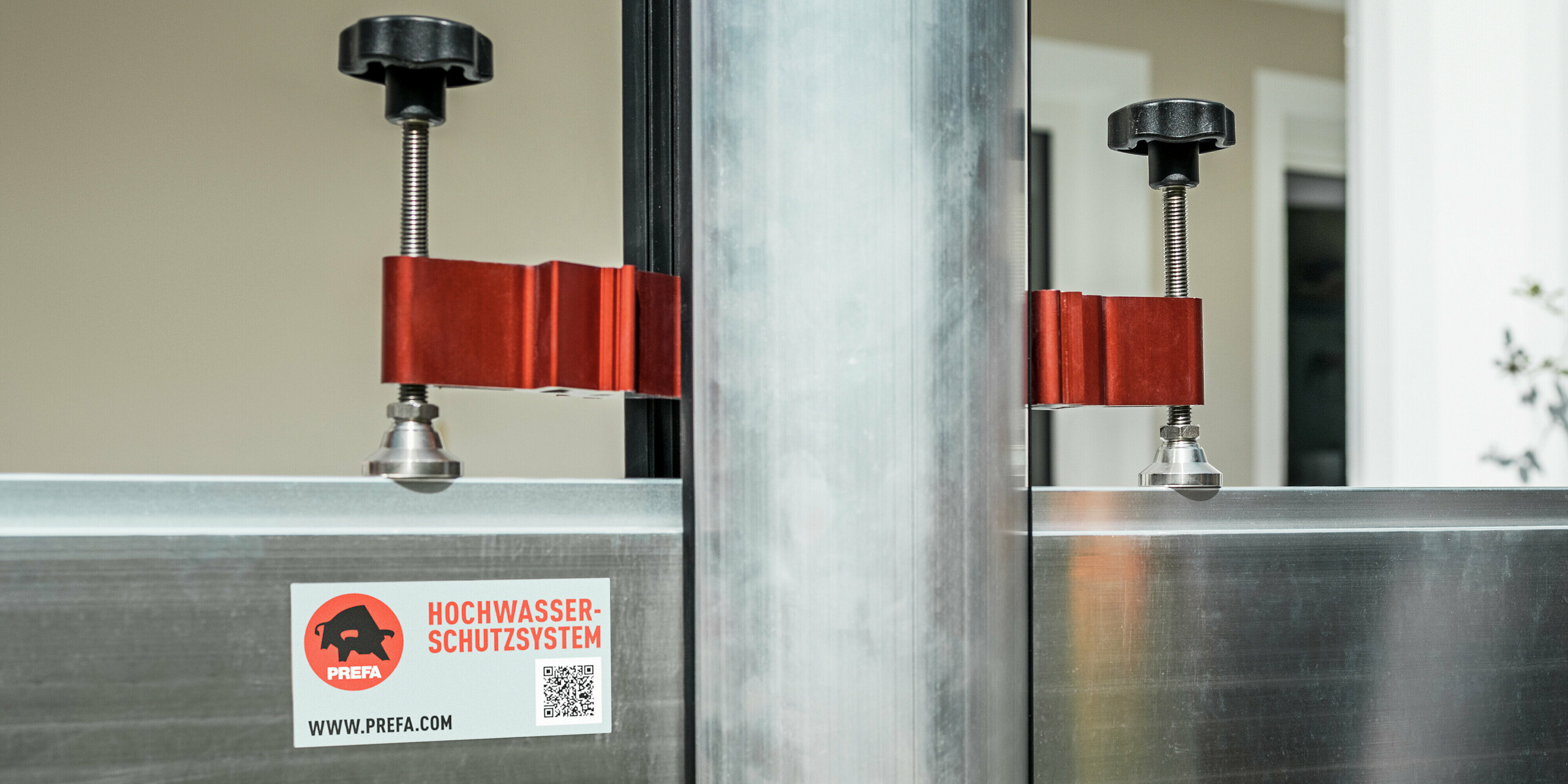 Nahaufnahme des PREFA Hochwasserschutzsystems mit Fokus auf die Spannvorrichtung zur Befestigung der Dammbalken. Gut zu erkennen sind die robusten Aluminiumprofile, die roten Spannelemente aus Metall und die schwarzen Handverschraubungen, die eine schnelle und sichere Montage ermöglichen. Das Bild zeigt die hochwertige Verarbeitung und Funktionalität des Systems - ein zentraler Baustein für den effektiven Hochwasserschutz von Gebäuden und Grundstücken.