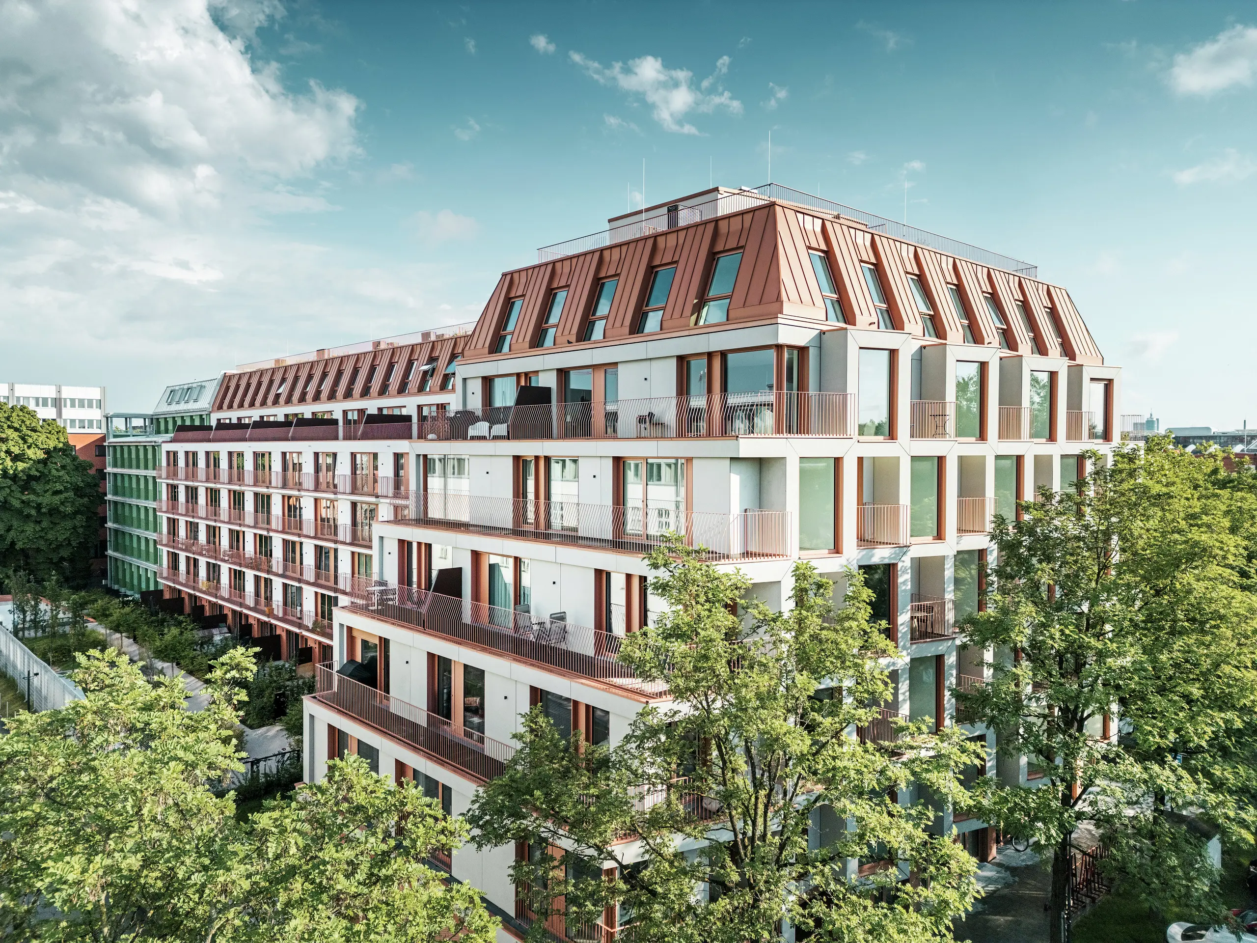 Overall view of the Van B urban residential project in Munich with its clear architecture, green outdoor areas and striking PREFALZ aluminium roof in IGP Orange HDP from PREFA. The durable and low-maintenance roofing material forms the connecting design element of the building and ensures a long-lasting, aesthetic solution in an urban space. Generous window areas, balconies and well-thought-out floor plans offer modern living comfort with high design quality.