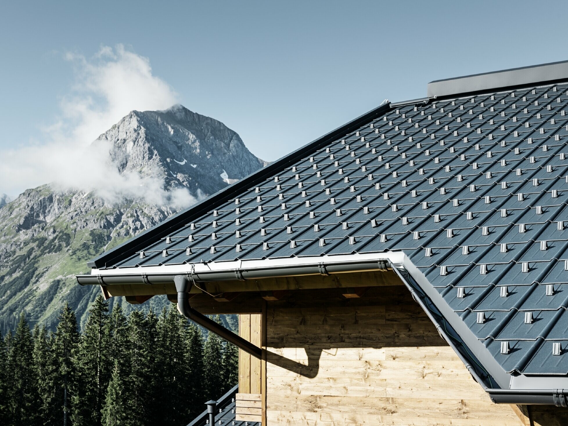 PREFA Dachplatte im alpinen Einsatz - Dachplatte in Anthrazit verlegt auf Hütte in Lech am Arlberg mit Berge im Hintergrund; PREFA Dachrinne in Anthrazit; Holzfassade