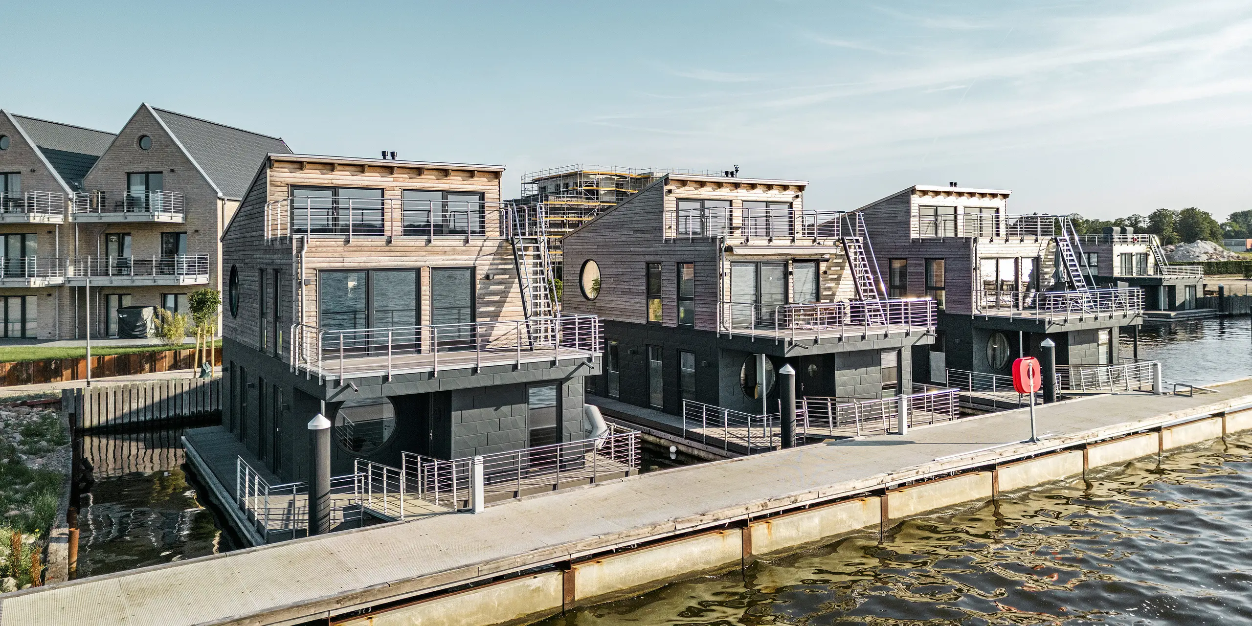 Einzigartige Wasserhaus-Komposition im Pionierhafen Schleswig, wo modernes Wohnen auf dem Wasser mit PREFA Siding.X in P.10 Anthrazit realisiert wird. Die Fassaden der Häuser kombinieren natürliche Holzelemente mit robusten Aluminiumpaneelen, die sich nahtlos in die Küstenlandschaft einfügen. Die Architektur reflektiert einen maritimen Lifestyle, begünstigt durch die direkte Lage am Wasser und unterstrichen durch die hochwertige, korrosionsbeständige Verkleidung, die Schutz und Ästhetik vereint.