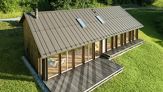 Modernes Holzhaus mit großzügiger Glasfassade und strapazierfähigem PREFALZ Aluminiumdach in P.10 Braun – ideal für Hanglagen wie in Vinje. Die hochwertige Dacheindeckung sorgt für maximale Wetterbeständigkeit, lange Lebensdauer und eine elegante Optik. Durch präzise Verlegung der Stehfalze und integrierte Dachfenster entsteht ein lichtdurchflutetes, nachhaltiges Wohnkonzept. Die Vogelperspektive zeigt die präzise Verarbeitung der Stehfalze aus Aluminium.