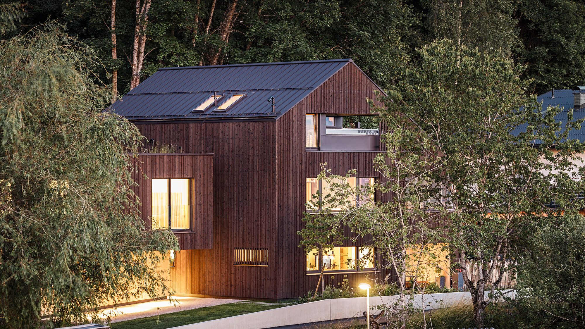 Evening shot of the multi-family home illuminated from the inside with its natural surroundings from a lateral perspective.