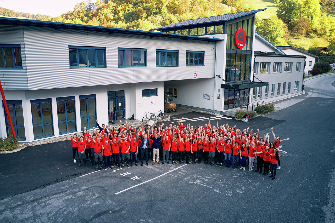 PREFA employees in red T-shirts in front of the PREFA headquarters in Marktl with cheering arms.