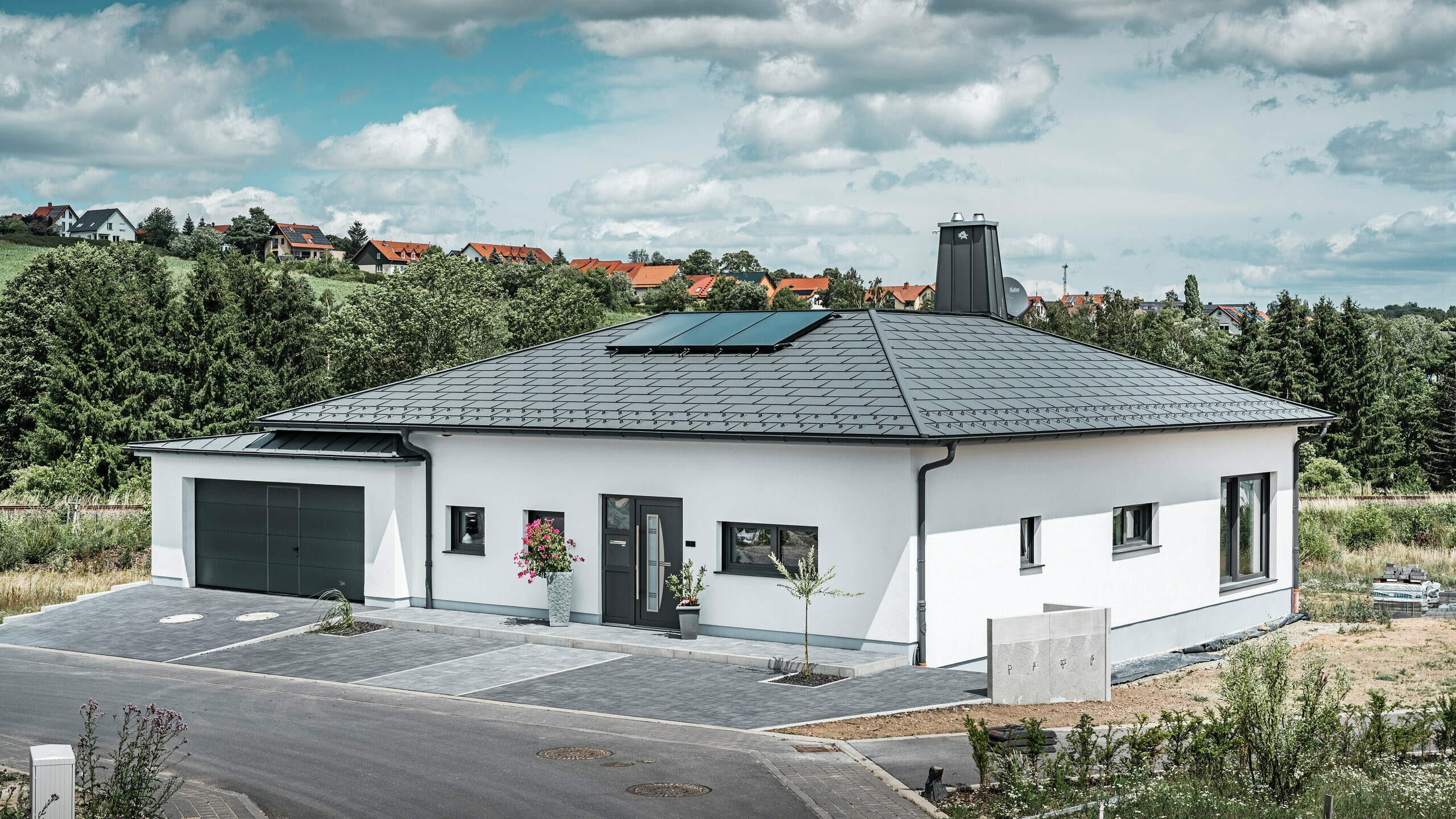 Elegant detached house in Ilmenau, Thuringia, with a high-quality PREFA roof in P.10 dark grey, fitted with robust R.16 roof tiles. The garage harmonises in colour and style thanks to the use of PREFALZ. The eye-catching features of the property are the functionally designed atrium and the chimney decorated with the PREFA bull, which underline PREFA's modern design and innovative roof technology. The solar panels fit seamlessly into the sustainable concept of the house. The well-thought-out overall concept of the bungalow is rounded off by the PREFA roof drainage system, which is also in dark grey.
