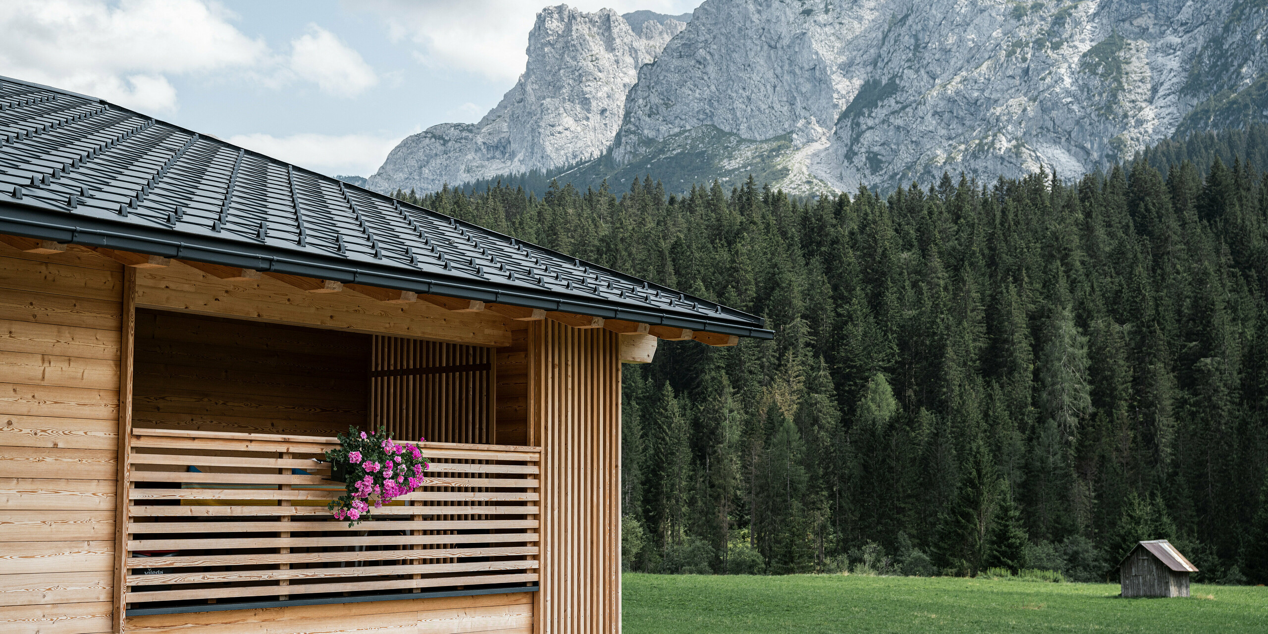 Casa Hoffe in Sappada, Norditalien, eingebettet in die Dolomiten, mit einem PREFA Dach aus Dachplatten R.16 in P.10 Schwarz. Die klaren Linien des Daches kontrastieren mit der natürlichen Holzfassade und der Loggia, die mit blühenden Pflanzen geschmückt ist. Dieses Bild vereint moderne Bautechnik mit traditioneller alpiner Wohnkultur, umgeben von beeindruckender Natur.