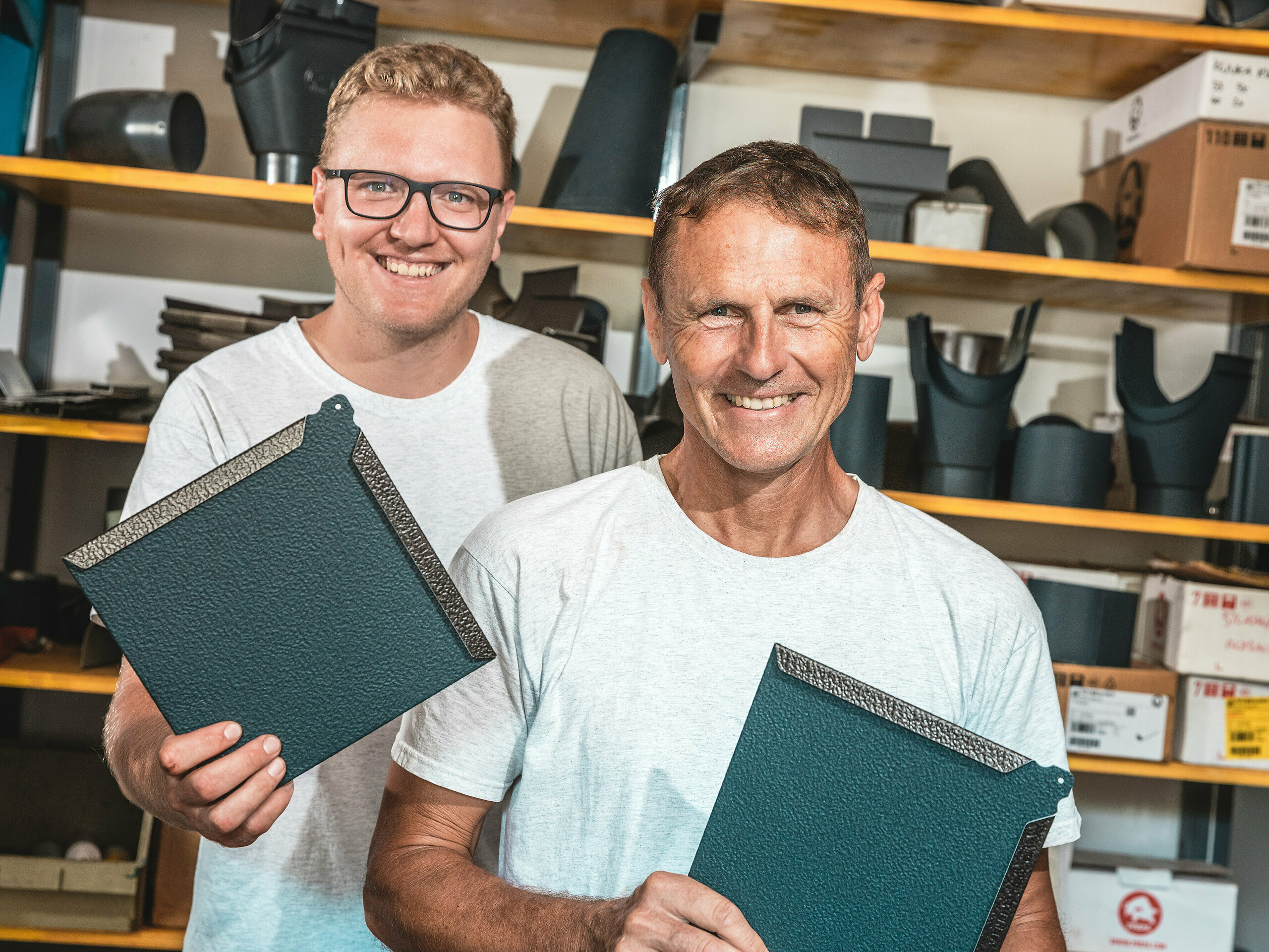 In einer Werkstatt präsentieren zwei Handwerker quadratische PREFA Wandrauten aus Aluminium in der Farbe P.10 Anthrazit. Der Mann im Vordergrund ist der slowenische Spenglermeister Janez Kralj. Hinter ihm steht ein junger Mitarbeiter aus seinem Team. Im Hintergrund sind Regale mit Bauteilen und Zubehör für Dach- und Fassadensysteme zu sehen. Die fein strukturierte Oberfläche der PREFA Aluminiumrauten zeugt von ihrer hohen Materialqualität, Langlebigkeit und präzisen Verarbeitung. Sie sind ideal für anspruchsvolle Architekturprojekte wie Tiny Houses, bei denen Widerstandsfähigkeit, geringes Gewicht und modernes Design gefragt sind.