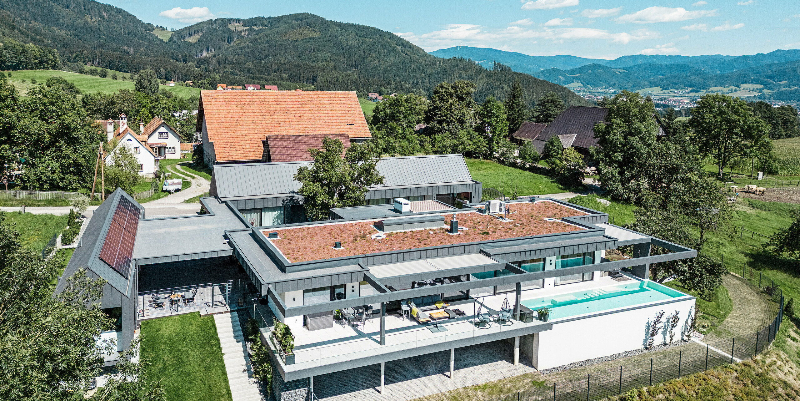 Drohnenaufnahme eines imposanten Einfamilienhauses mit landwirtschaftlich genutzten Gebäudeteilen, Terrasse und Swimmingpool in Pogier, Österreich. Umrahmt werden die dunkelgrauen Gebäude von einer atemberaubenden Berglandschaft. Das Objekt präsentiert sich mit einer PREFALZ Dach- und Fassadenbekleidung in dunkelgrau P.10, die die klaren, modernen Linien unterstreicht. Die Terrasse und der Swimmingpool bieten einen luxuriösen Außenwohnbereich mit Panoramablick auf die umliegende Landschaft. Die Kombination aus den modernen Bauelementen von PREFA und der idyllischen Lage schafft eine perfekte Symbiose aus zeitgemäßem Design und natürlicher Schönheit.