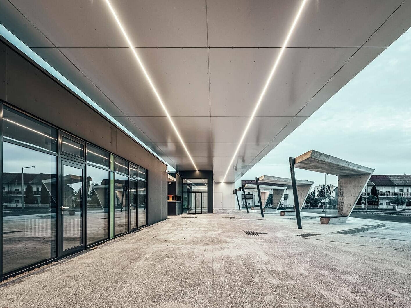 The railway station at dusk. Two LED light stripes are floating through the ceiling of the station. On the right side are several bus stops with a waiting area.