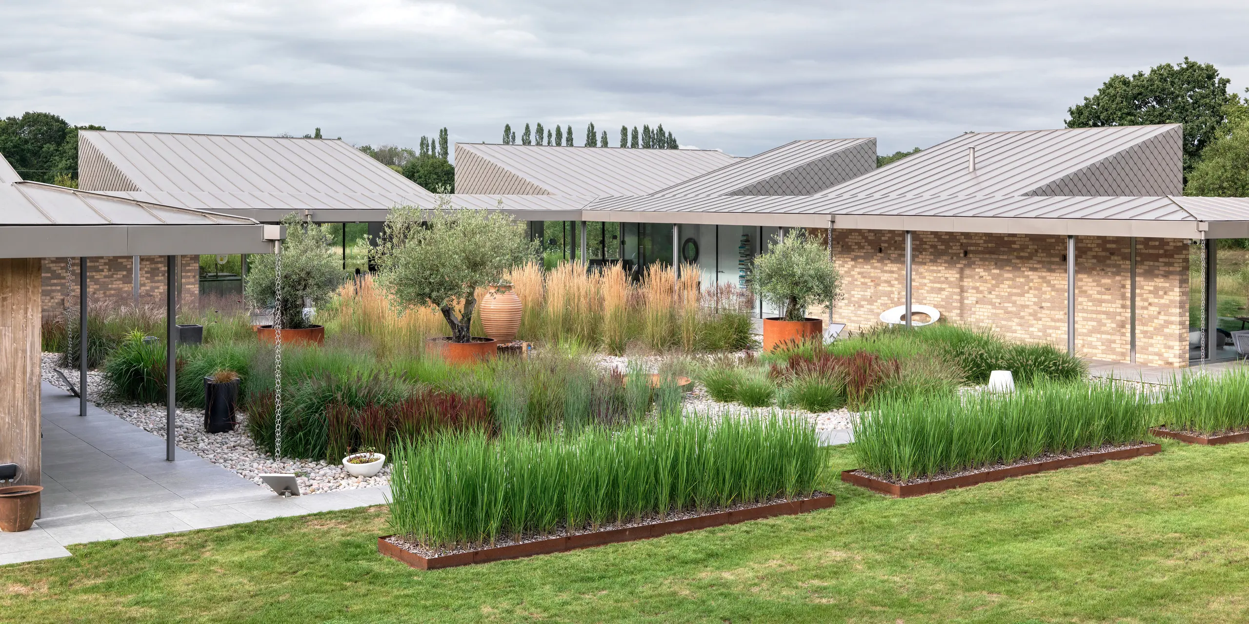 Spacious garden view of a modern bungalow in West Sussex with multi-sectioned standing seam roofs in P.10 bronze. The focal point is a natural courtyard with grasses, olive trees and terracotta elements, surrounded by covered walkways with filigree outlets. The clear roof lines made of durable PREFA aluminium emphasise the architectural structure while offering maximum weather resistance and low maintenance.