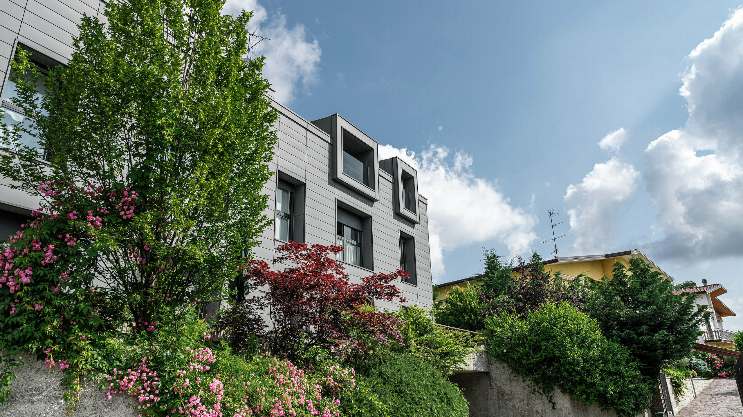Modern residential complex in Faloppio, Italy, with a stylish façade clad in PREFA siding. The cantilevered windows provide a unique architectural feature, while the surrounding plants and flowers emphasise the natural beauty of the property. The durable aluminium cladding from PREFA provides an aesthetic and functional solution in all weather conditions.