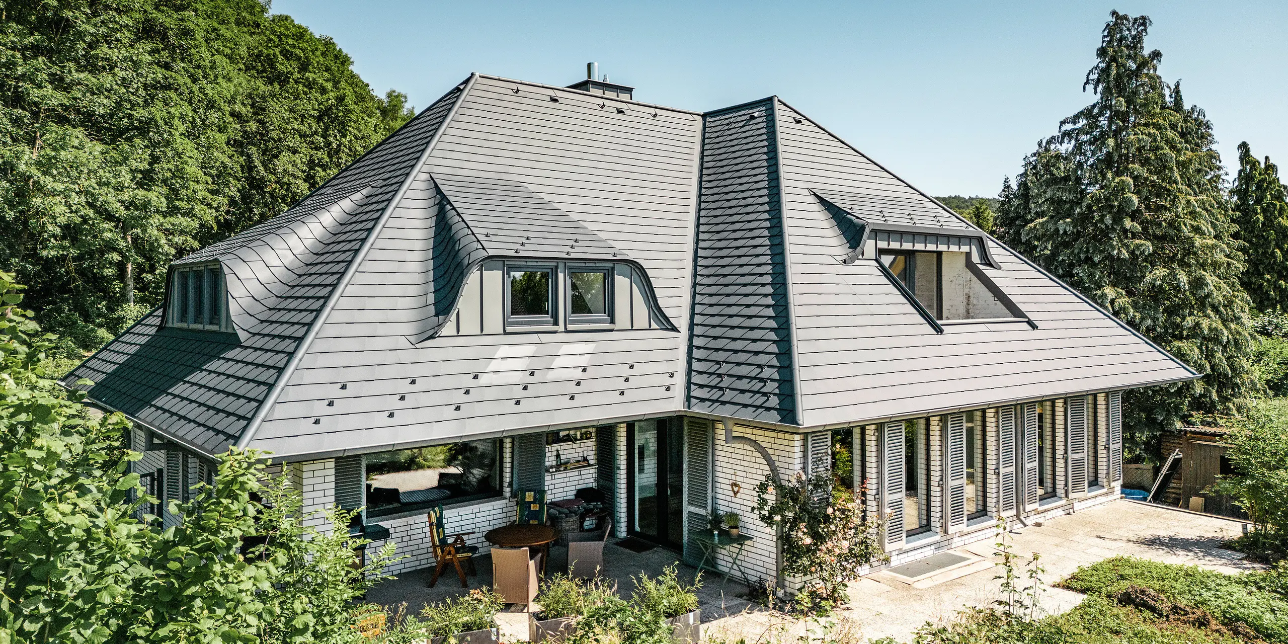 Modern detached house in Einbeck, Germany, with PREFA shingles in P.10 anthracite. The durable PREFA aluminium shingles are renowned for their quality and longevity. The roof design of the building emphasises the individual architecture with dormer windows, which are highlighted by the flexibility of the PREFA shingles. The roof drainage in the form of PREFA downpipes and half-round gutters is also in anthracite. The detached house is surrounded by a spacious and well-kept garden and a terrace. Behind the house is a forest which, together with the blue sky, creates a natural atmosphere.