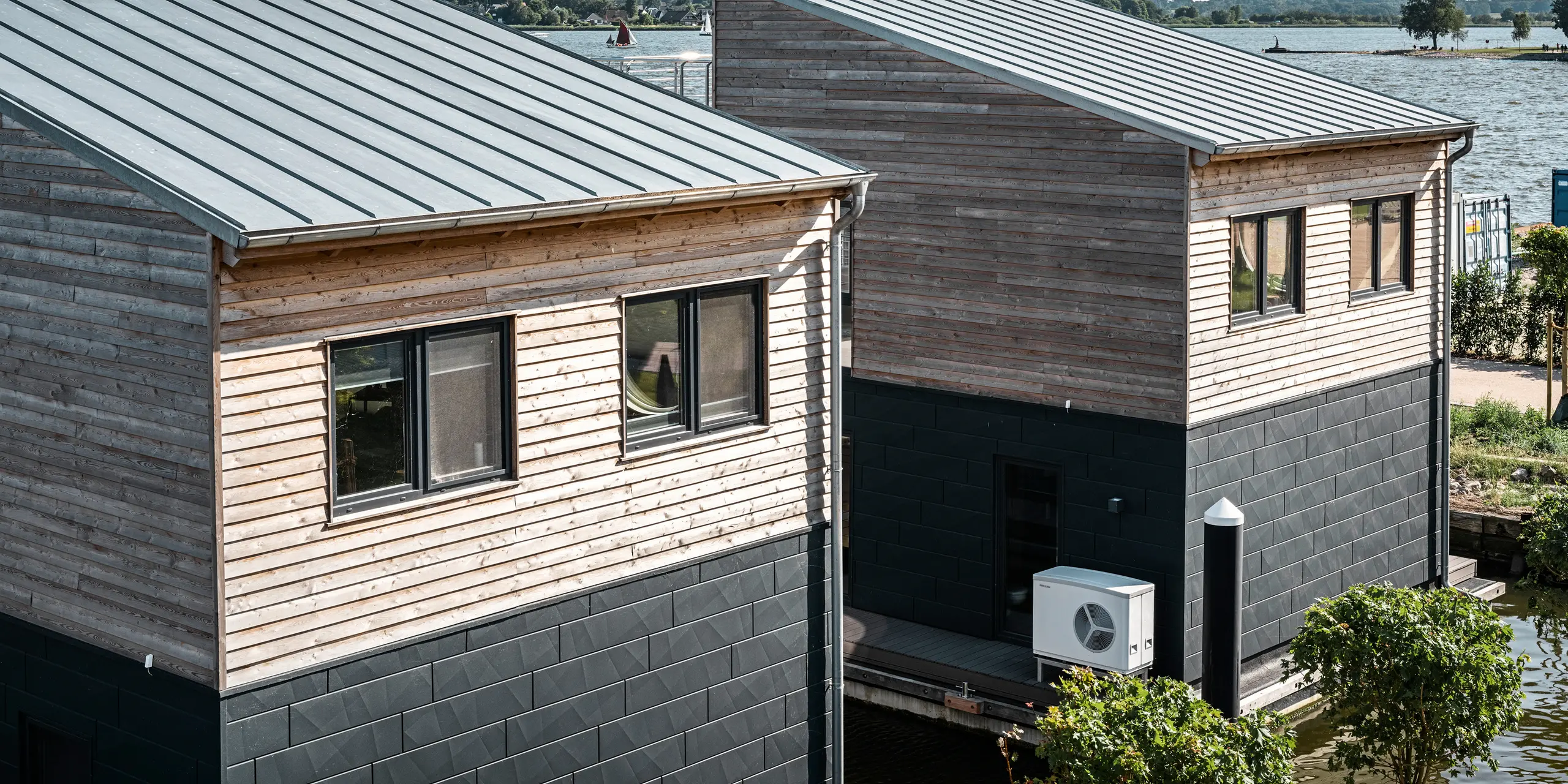 Drohnenaufnahme von zwei Wasserhäusern im Pionierhafen Schleswig, die eine attraktive Kombination aus naturbelassenen Holzoberflächen und dunklen PREFA Siding.X Aluminiumpaneelen in P.10 Anthrazit aufweisen. Die Architektur integriert moderne Haustechnik, wie die sichtbare Wärmepumpe, die für eine umweltfreundliche Klimaregulierung sorgt. Die Häuser sind durch ihre moderne Bauweise und robuste Materialwahl ideal an das Leben am Wasser angepasst, wodurch sie eine alternative Wohnoption darstellen.