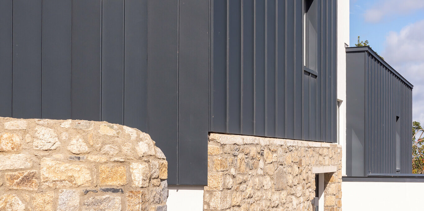The photo shows the modern façade of a terraced house in Watson Place with clean lines and a striking combination of natural stone walls and dark-looking PREFALZ standing seam cladding in P.10 anthracite. The standing folds of the aluminium profile create vertical accents and give the building an elegant structure. The weather-resistant PREFA aluminium provides lasting protection against moisture and wind, while the lightweight construction allows for flexible design. The interplay of robust natural stone and high-quality aluminium creates a timeless yet dynamic overall impression.