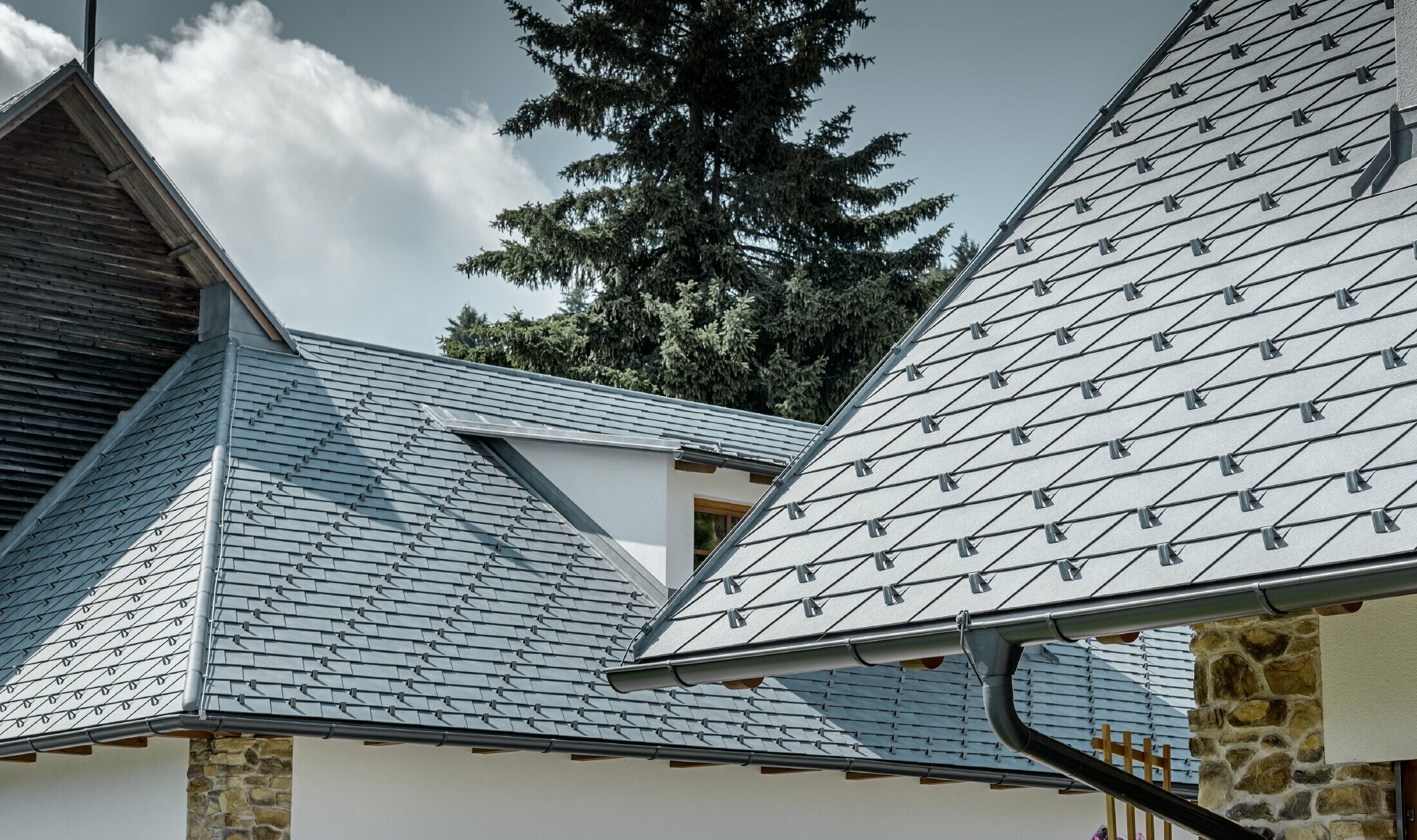 Close up of a PREFA aluminium roof cover; roof shingles in stone grey with the PREFA aluminium gutter in anthracite; an eyebrow dormer with standing seam roof can be seen in the background. The façade is white with integrated stone elements.
