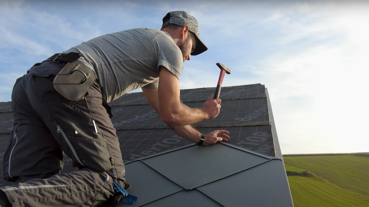Installer in action: The owner of the company ‘Der Blechmann’ uses a hammer to precisely install PREFA rhomboid roof tiles 44 × 44. The precision of the installation can be seen in the rhomboid roof tiles already installed at the bottom of the image. The man is wearing a cap and kneeling on the roof with his back to the camera. The clear sky and green landscape can be seen in the background. The harmonious background emphasises the symbolism of the image: sustainability and durability in craftsmanship with high-quality PREFA aluminium products.