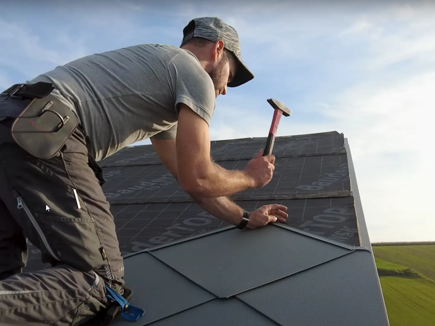 Installer in action: The owner of the company ‘Der Blechmann’ uses a hammer to precisely install PREFA rhomboid roof tiles 44 × 44. The precision of the installation can be seen in the rhomboid roof tiles already installed at the bottom of the image. The man is wearing a cap and kneeling on the roof with his back to the camera. The clear sky and green landscape can be seen in the background. The harmonious background emphasises the symbolism of the image: sustainability and durability in craftsmanship with high-quality PREFA aluminium products.