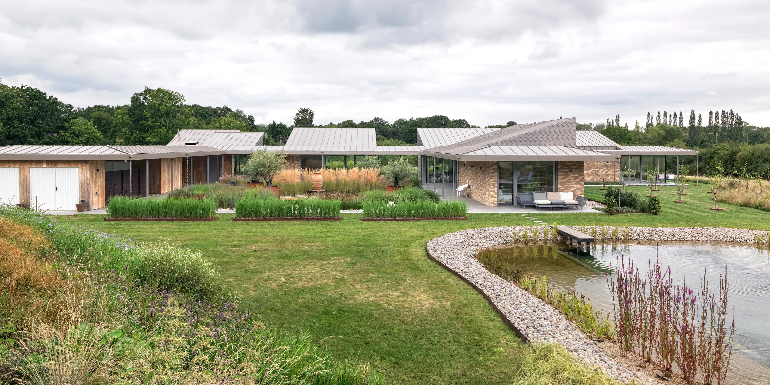 Wide-angle view of the architecturally sophisticated residential building in West Sussex with a large standing seam aluminium roof in P.10 bronze and harmoniously integrated PREFA rhomboid façade tiles in selected areas of the façade.The modern building extends flat and spacious across the property, nestled in natural landscaping with a landscaped natural pool in the foreground. The robust PREFA products support the sustainable building concept and ensure a uniform, long-lasting appearance.