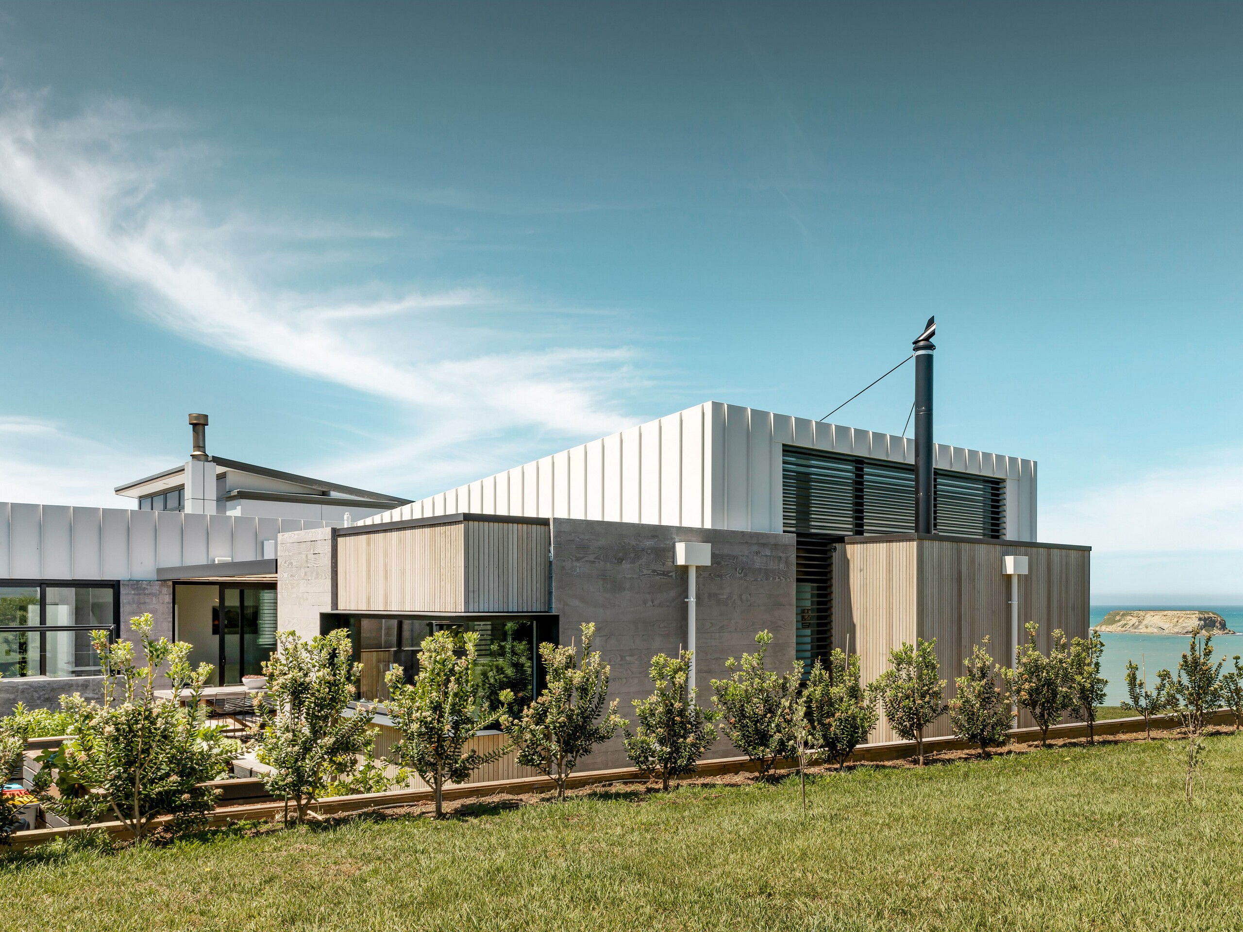 The Whitecaps House and its combination of the materials aluminium, wood and concrete from a side view. There is greenery in the front and Motunau Island and the Pacific can be seen in the background.