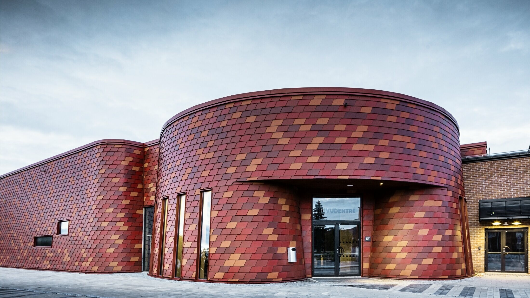 Ice rink with a flat roof and curved façade, the façade is clad with wall shingles made of aluminum from PREFA in different shades of red - oxide red, brick red, red-brown, brown-red