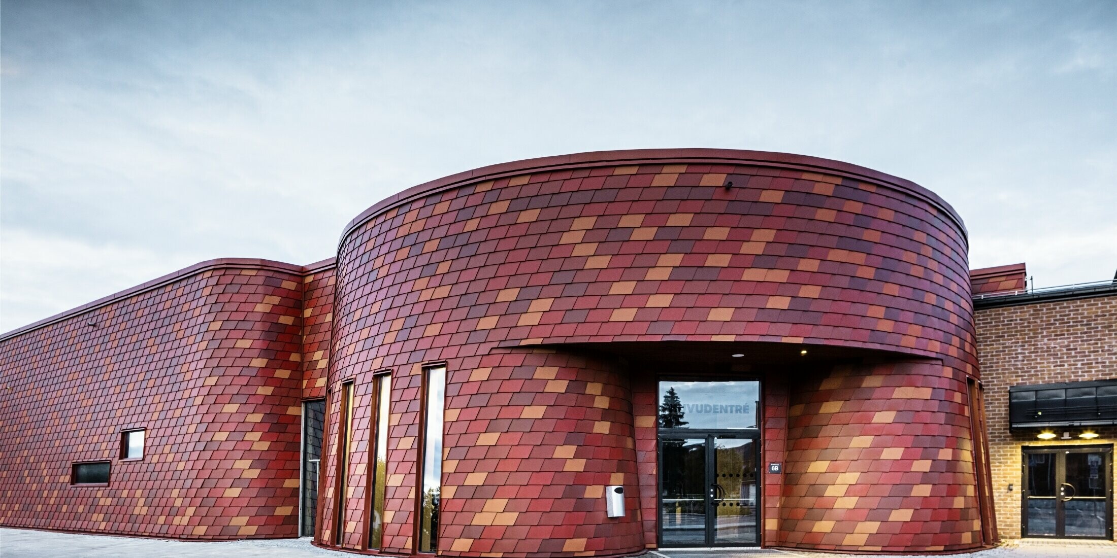 Ice rink with a flat roof and curved façade, the façade is clad with wall shingles made of aluminum from PREFA in different shades of red - oxide red, brick red, red-brown, brown-red
