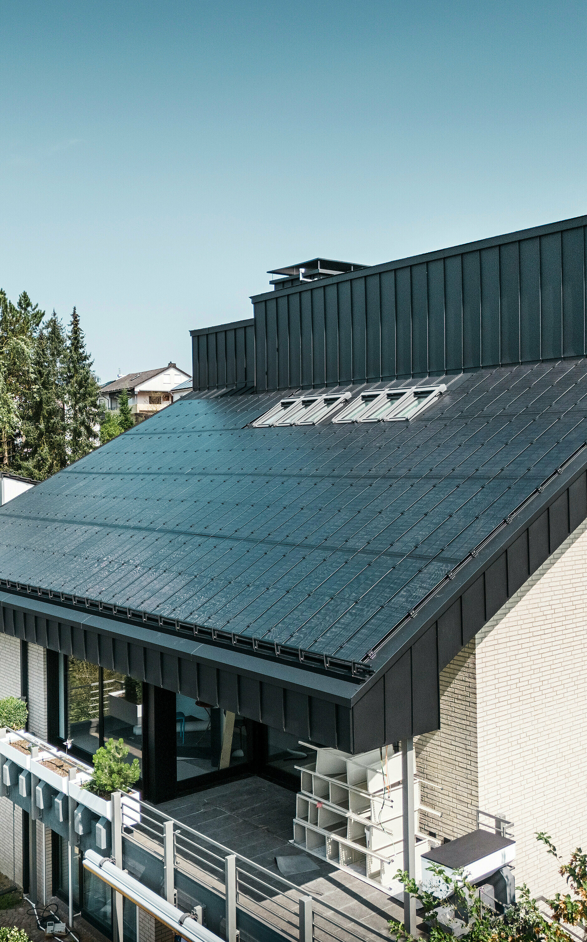 Moderne Architektur eines Einfamilienhauses in Meschede, Deutschland, mit einem PREFALZ Aluminiumdach in P.10 Schwarz und integrierten Glas/Glas PV-Modulen. Das versetzte Pultdach verbindet zeitloses Design mit nachhaltiger Photovoltaik-Technologie. Die innovativen PREFALZ Solarmodule liefern eine Leistung von 18 kWp. Die rahmenlosen Module und die elegante Aluminiumverkleidung von Fassade und Dach bilden eine ästhetische und funktionale Einheit, die sich perfekt in das grüne Wohnumfeld einfügt.