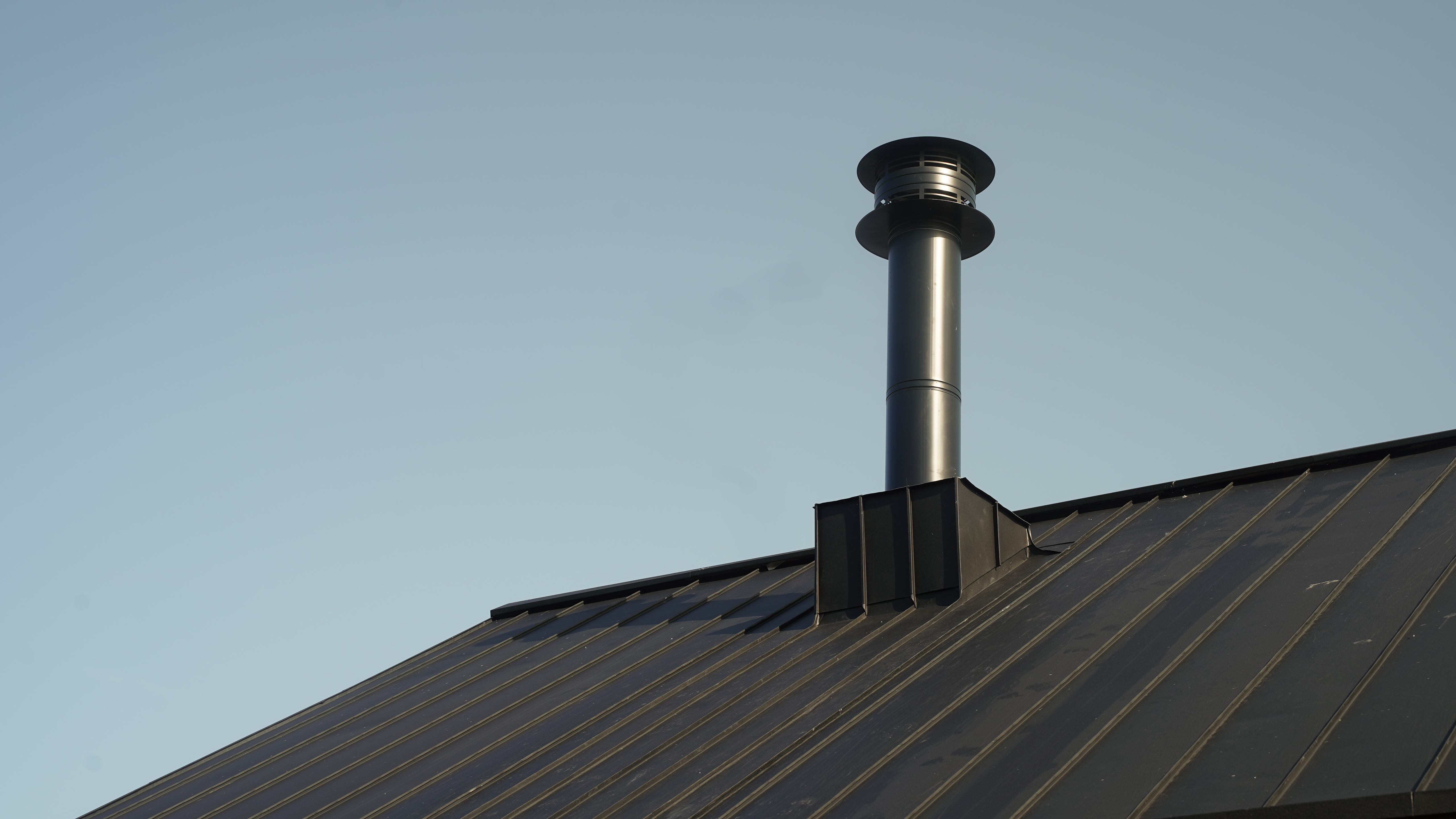 Close-up of a PREFALZ roof system in P.10 black on the "Golf it!" complex in Glasgow, Scotland, with a striking, modern chimney. The chimney rises elegantly above the precisely laid, dark standing seam sheets, which are known for their durability and robustness. This image underlines the combination of functionality and design that PREFA aluminium products offer.