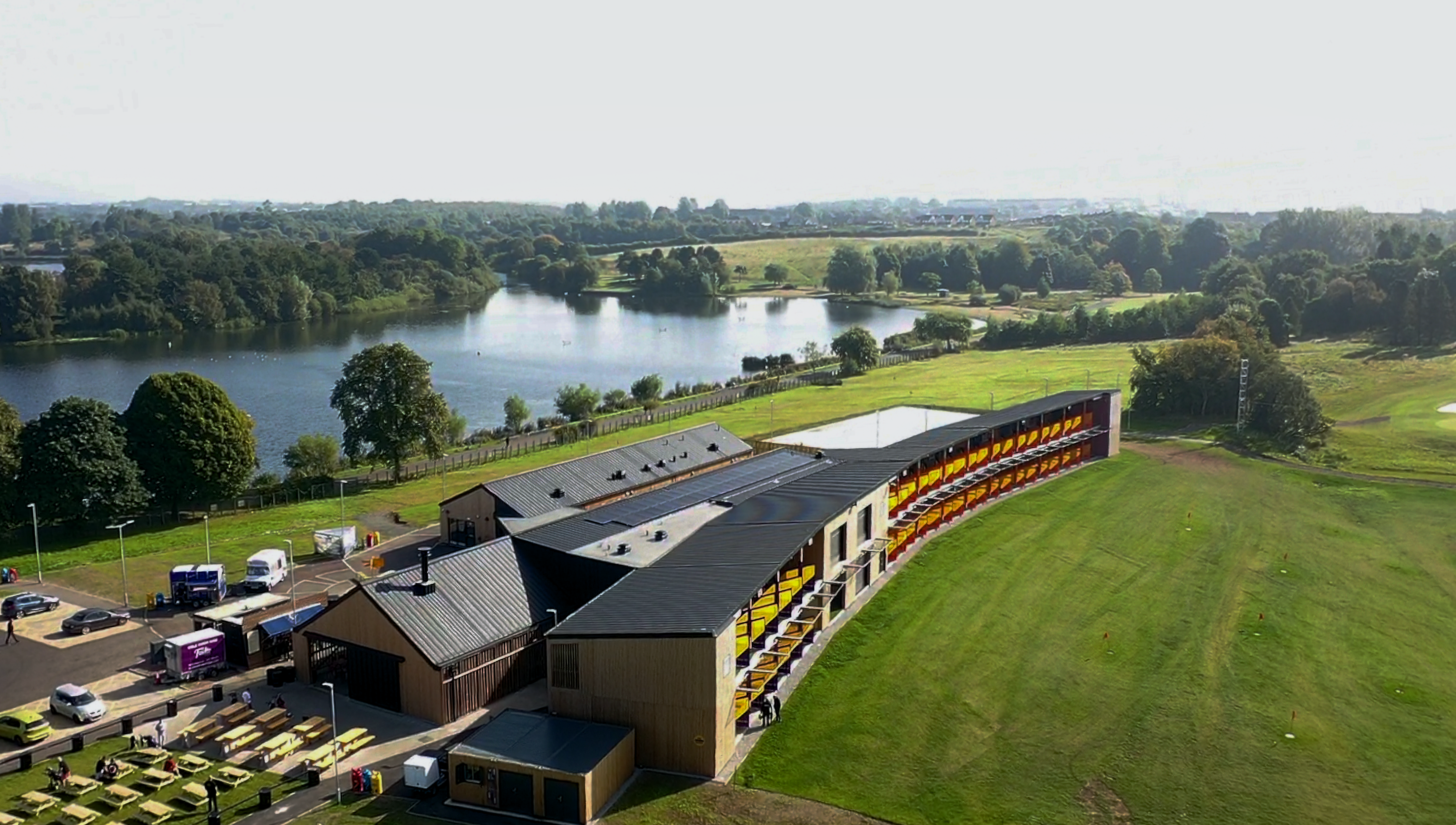 Drone shot of the 'Golf it!' driving range and golf venue in Glasgow, Scotland, with a PREFALZ roof system in P.10 black. The dark roof design creates an elegant contrast to the wooden façade of the building complex. The structure blends seamlessly into the surrounding landscape, with a clear view of the neighbouring lake and the lush greenery of the golf course. The roofs of the individual building components with their uniform, black Prefa aluminium cladding stand out against the natural backdrop and demonstrate the harmonious combination of modern architecture and natural beauty.