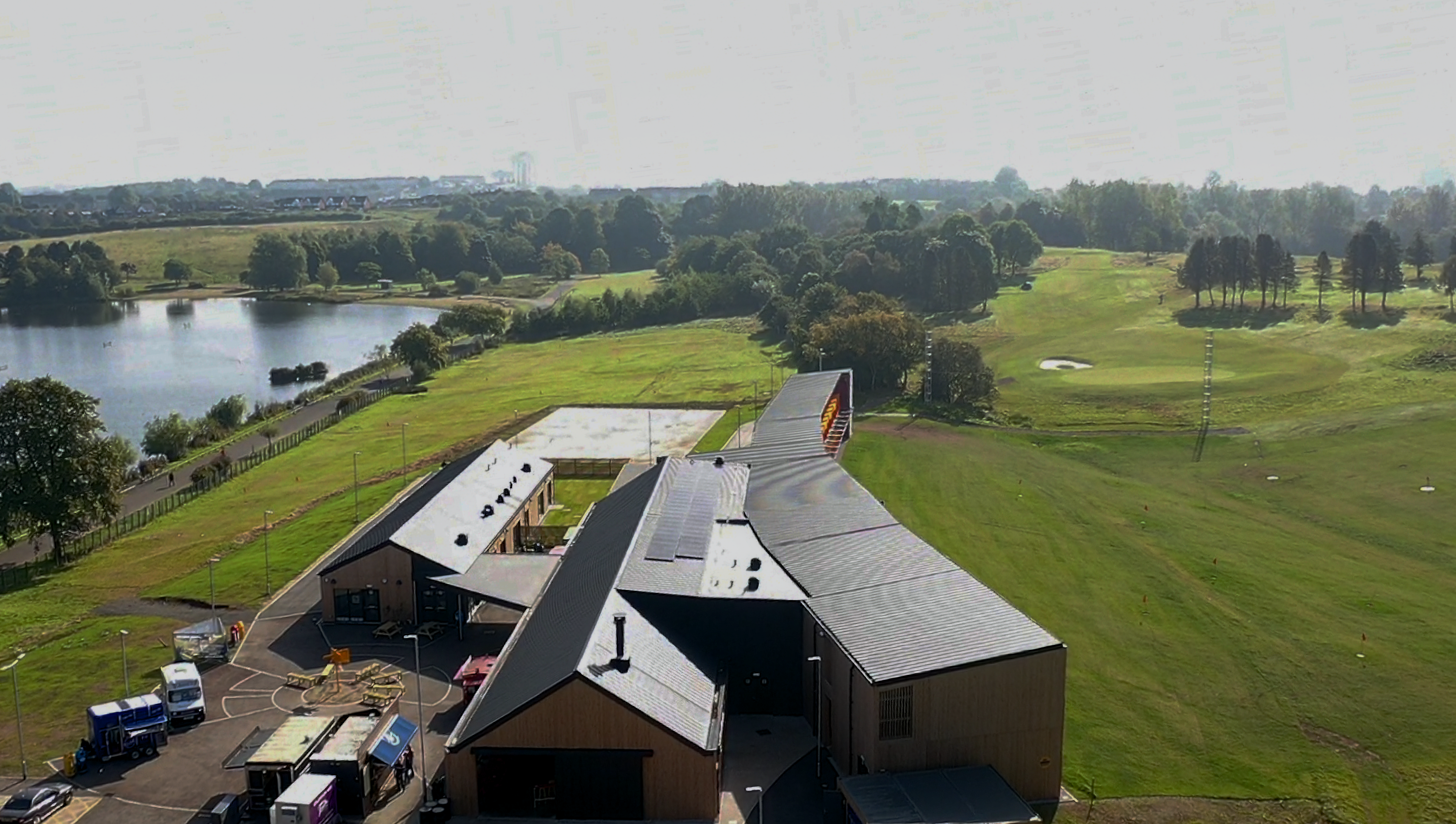 Aerial view of the 'Golf it! golf course in Glasgow, which is set in a stunning landscape with a lake and an extensive golf course. The buildings are clad with PREFALZ in P.10 Black. This view demonstrates the adaptability and aesthetic quality of PREFA aluminium products in a natural environment.
